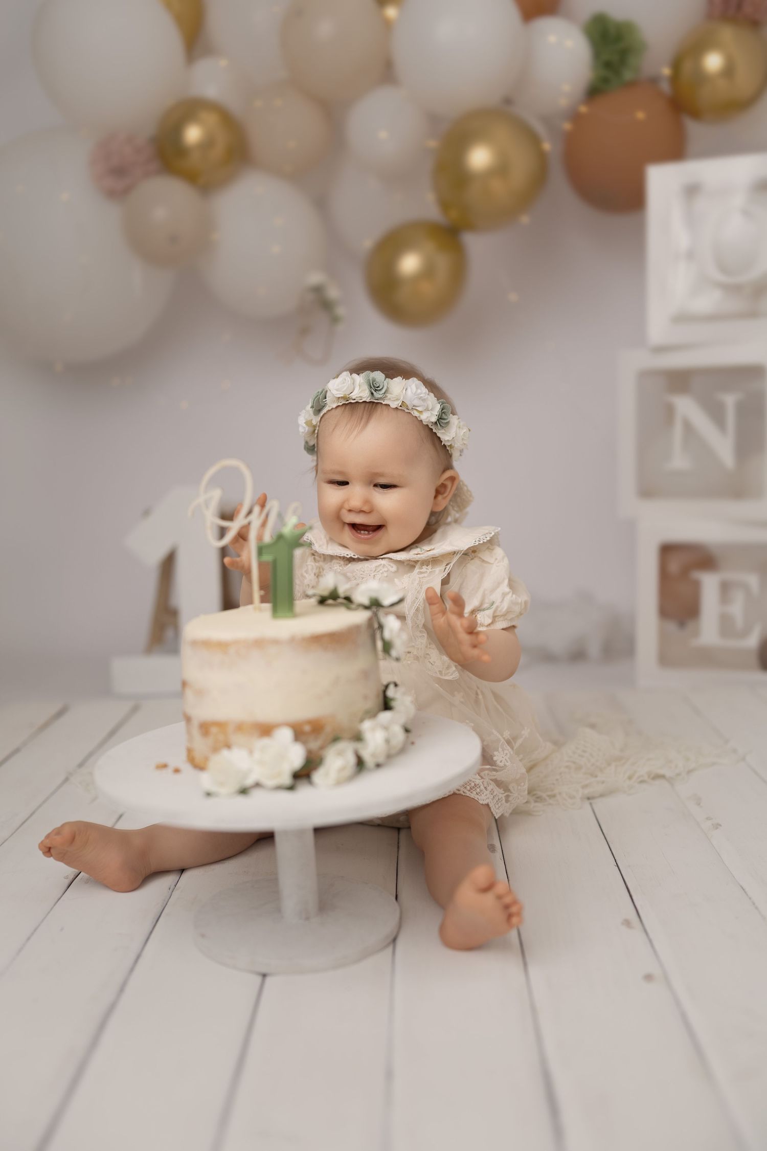 Baby in white dress and floral crown reaching for birthday cake during cake smash photoshoot.