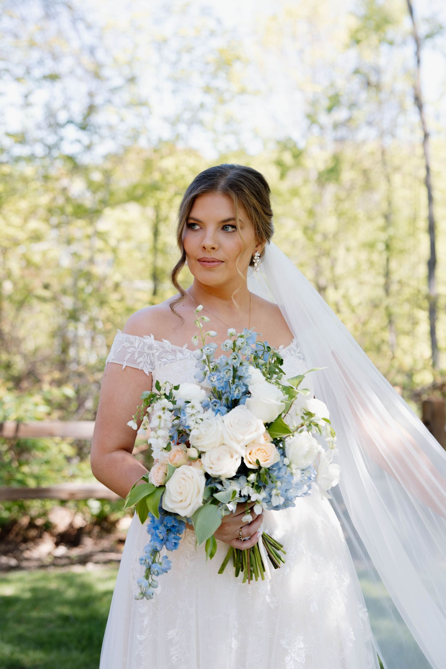 Romantic outdoor wedding portrait with a white off-shoulder gown and beautiful blue and white bouquet with trailing veil.