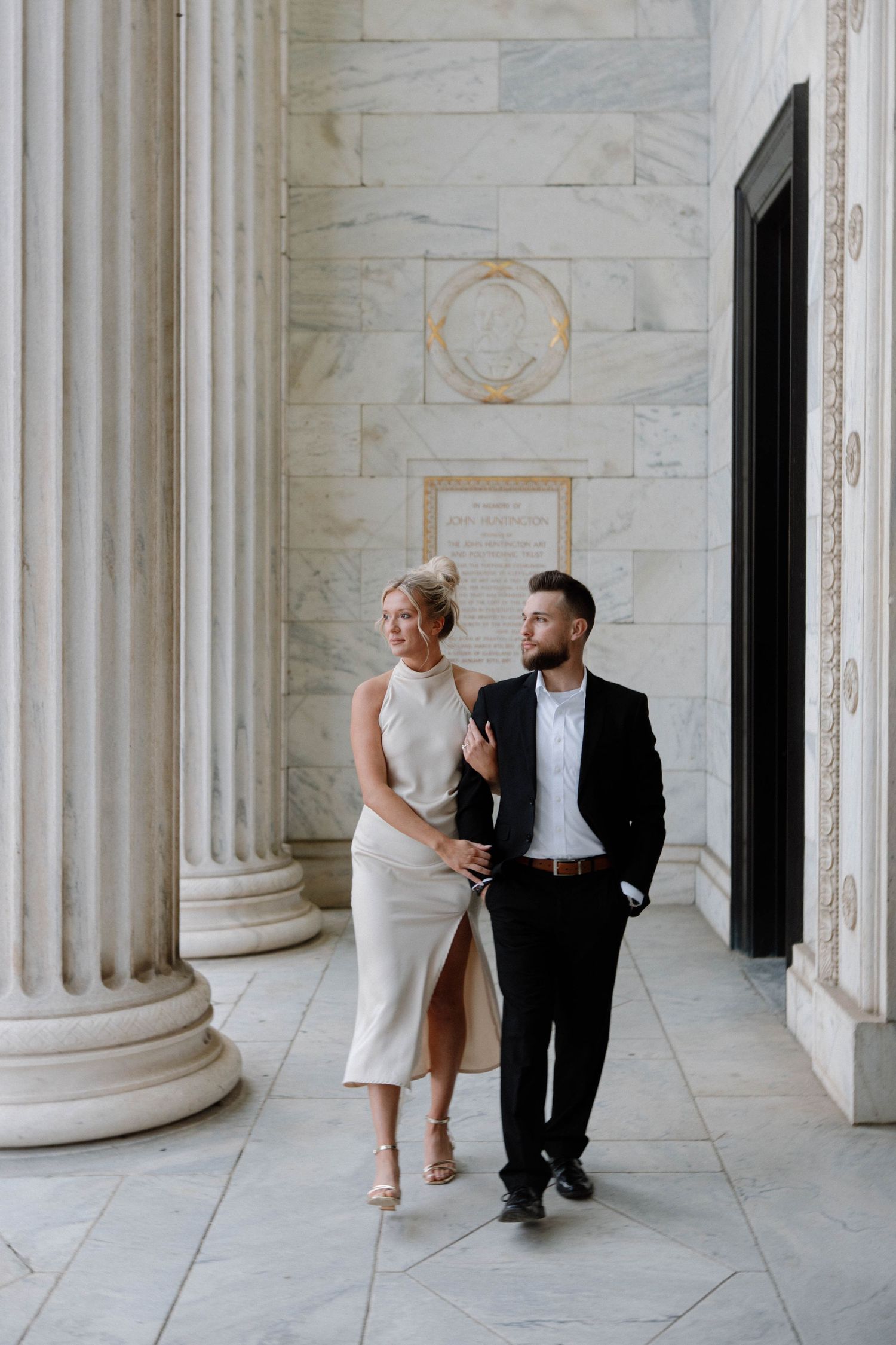 Elegant couple in formal attire walking together between white marble columns in a grand architectural setting. Cleveland, OH