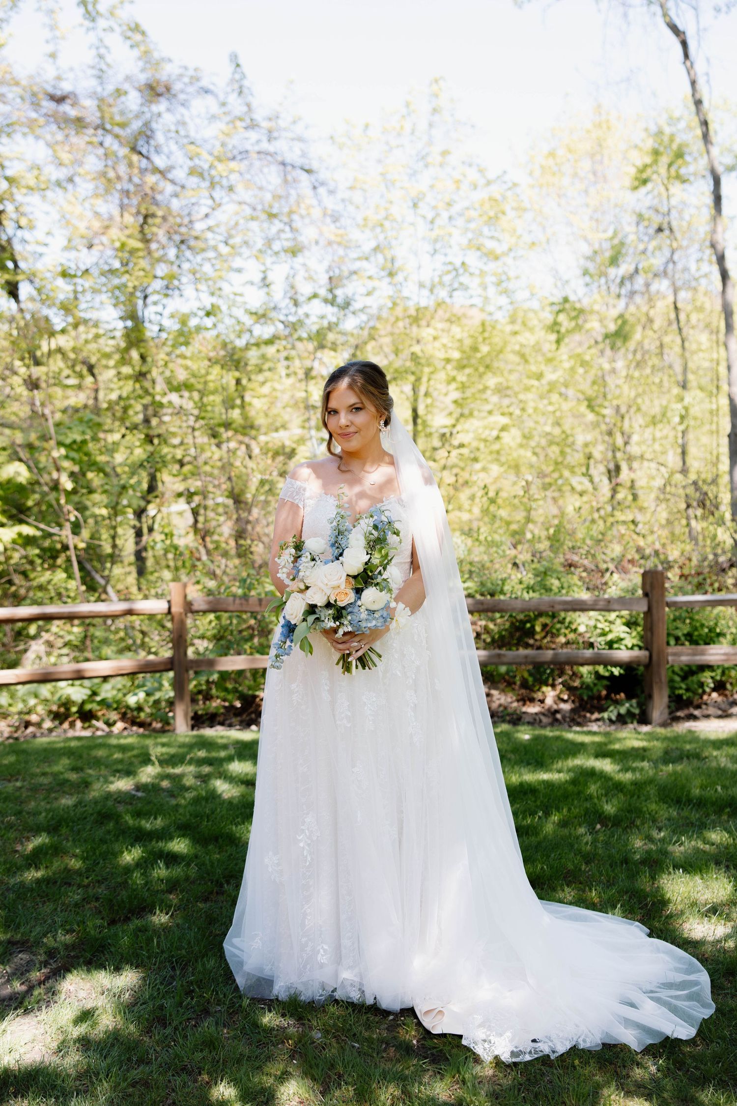 A bride in a flowing white wedding gown and veil poses with her bouquet in front of a rustic wooden fence outdoors.