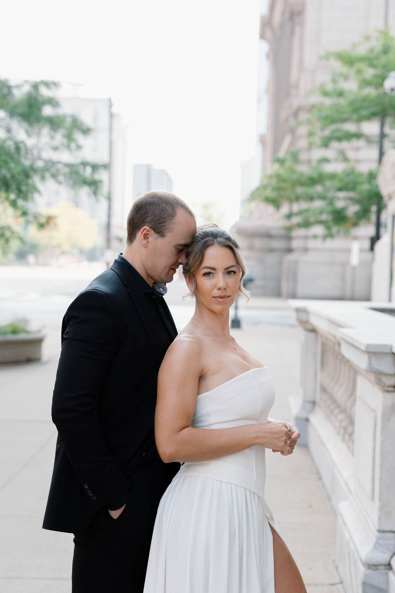 A couple in formal wedding attire pose together on an elegant city street with classic architecture in the background.