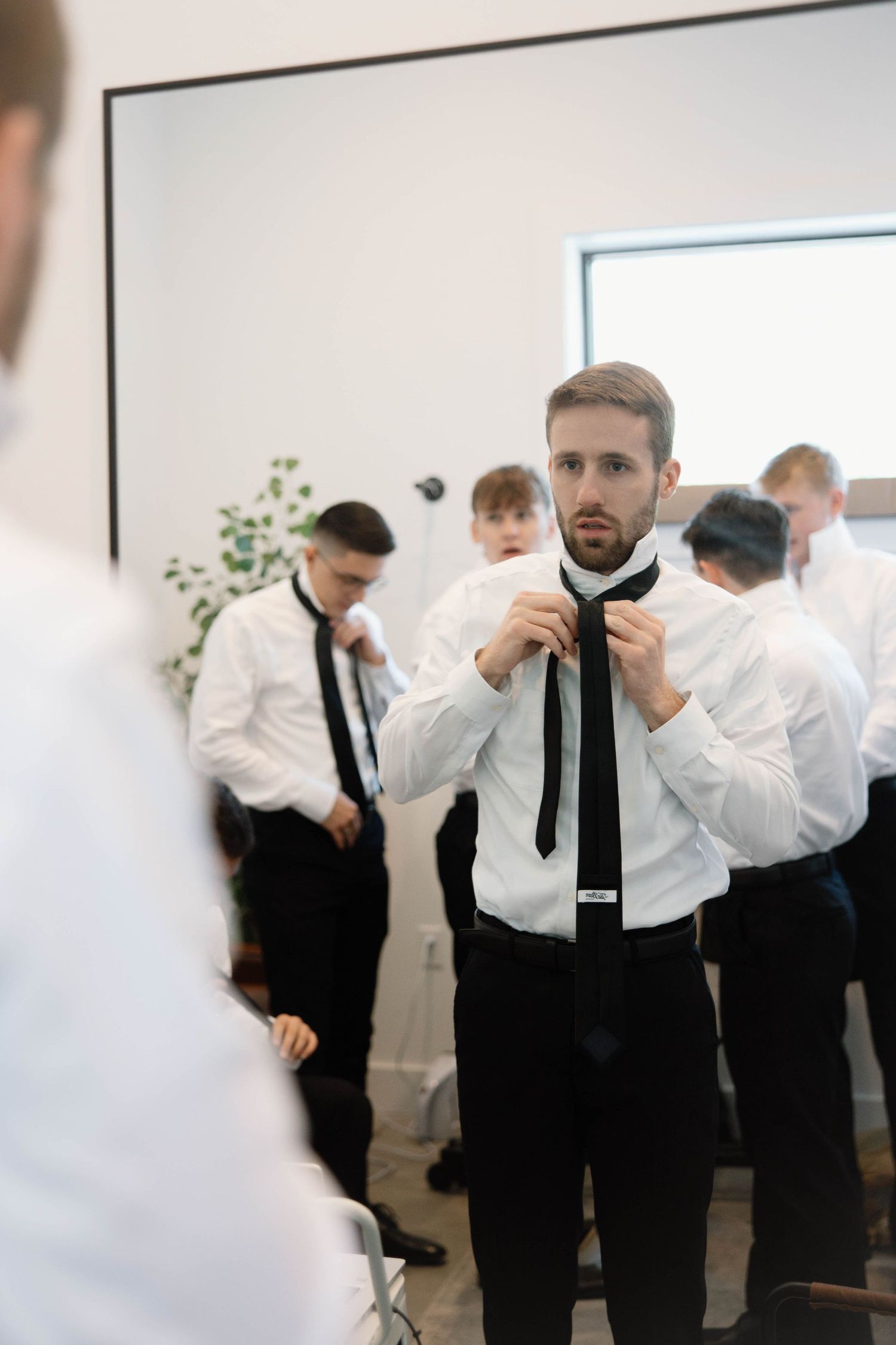 Well-dressed groomsmen in white shirts and black ties prepare for a wedding ceremony in a bright room.