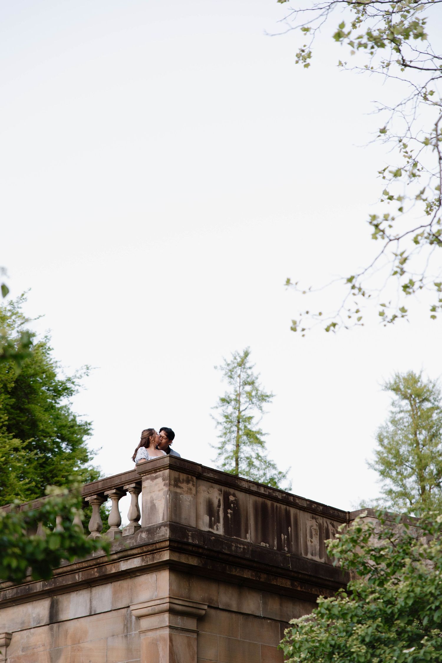Two people share a romantic moment on a stone balustrade overlooking trees and sky in an architectural garden setting.