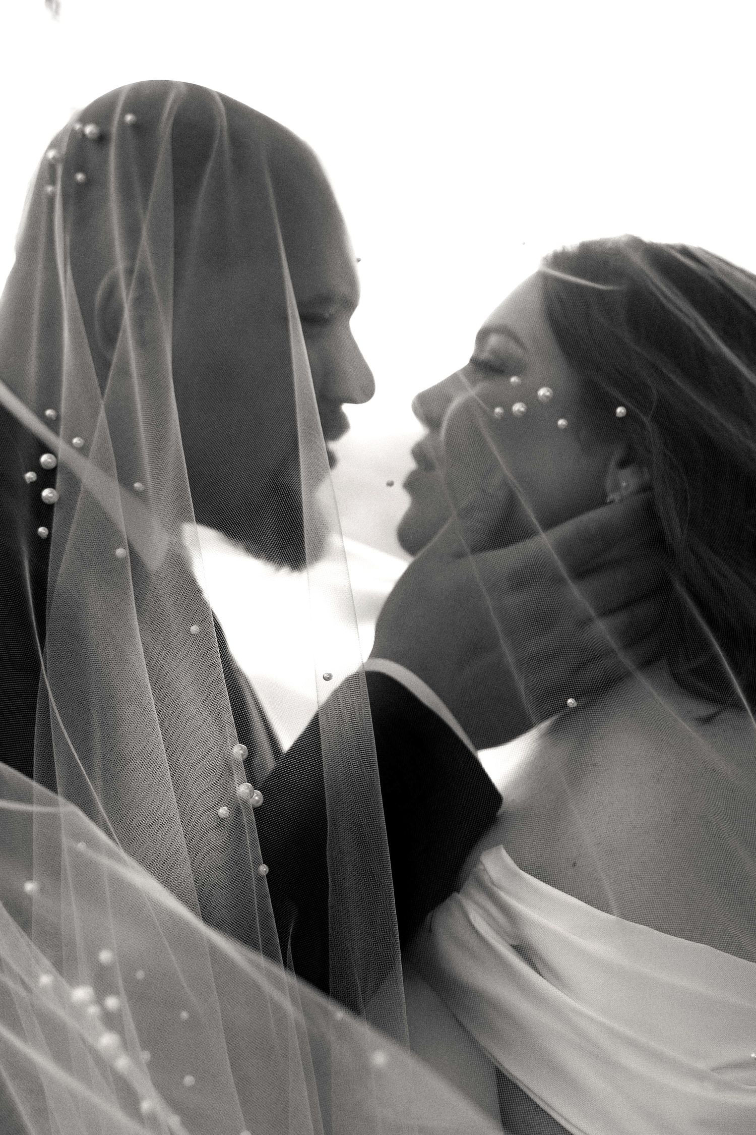 A black and white artistic wedding photo showing a couple in close embrace under delicate flowing veils.