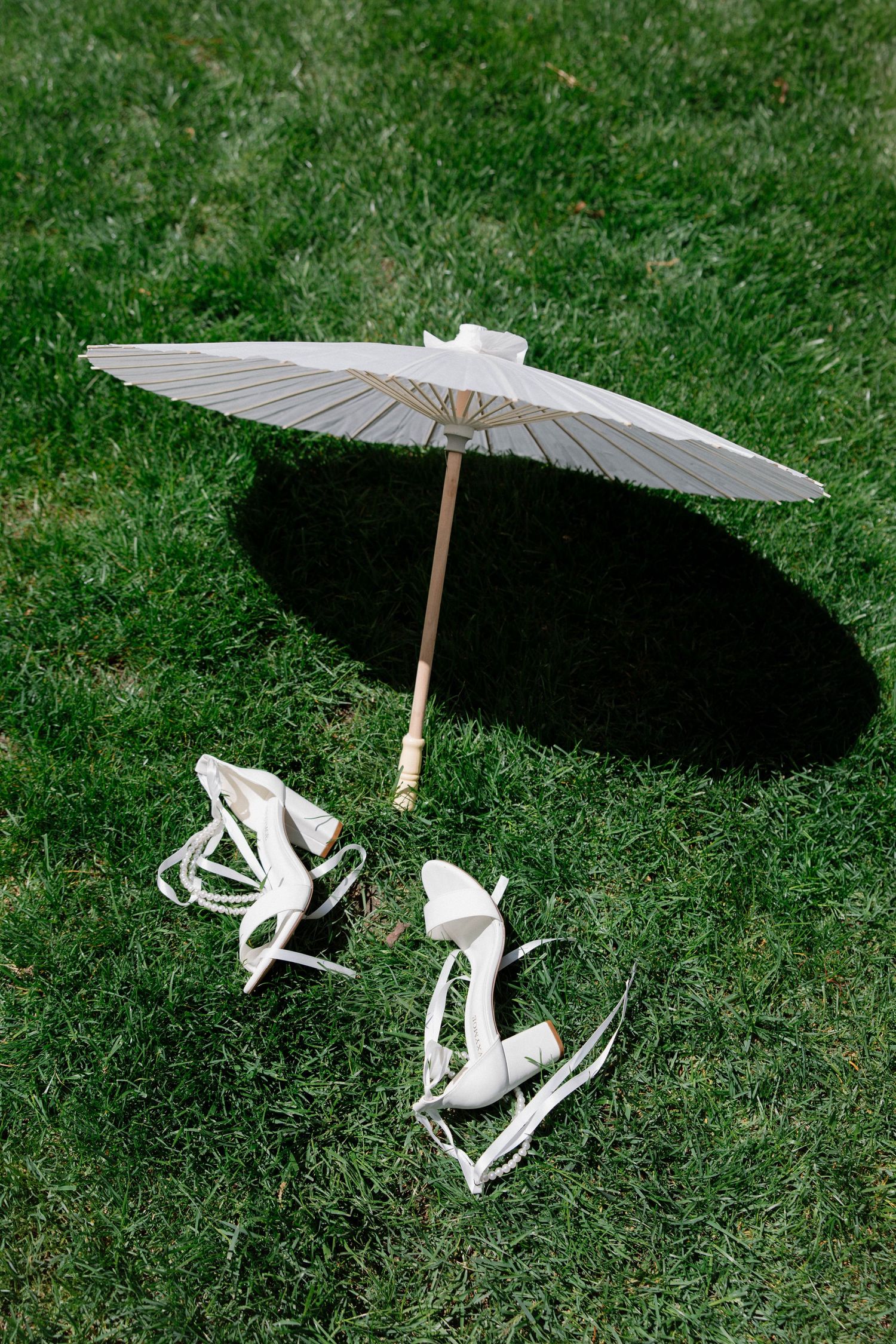 A white paper parasol and white bridal shoes arranged on green grass creating an elegant wedding detail setup.