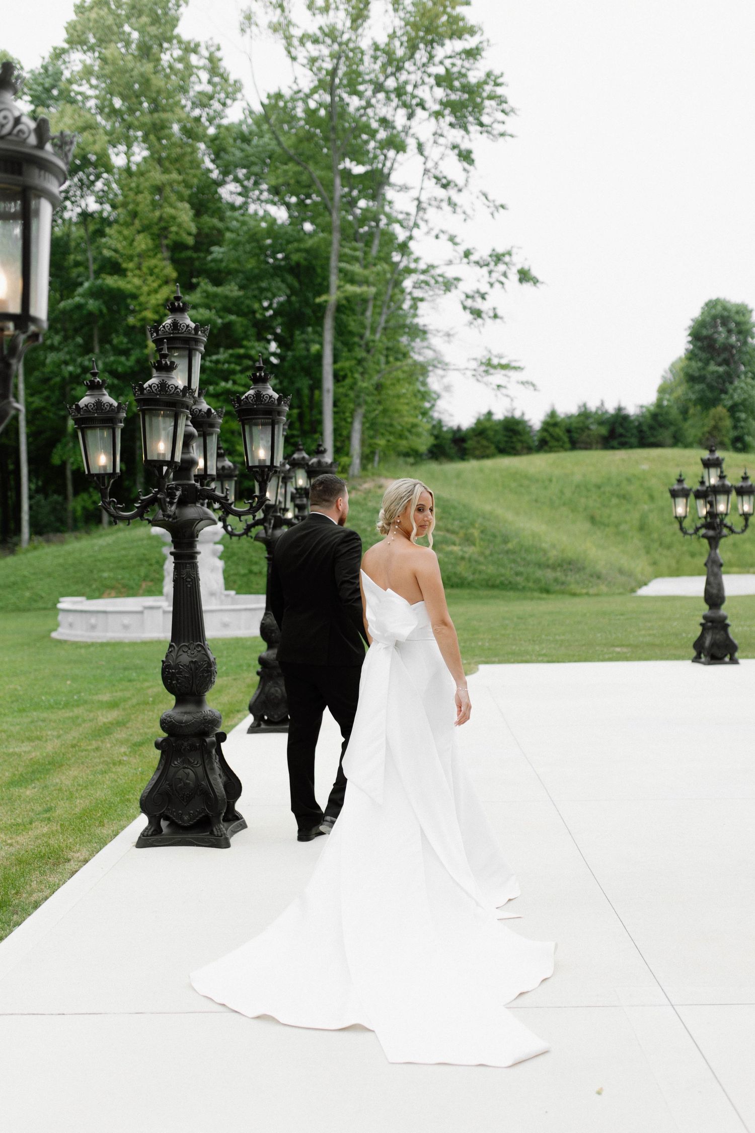 Bride in flowing white dress walks with groom along path with ornate black lampposts. Cleveland, OH
