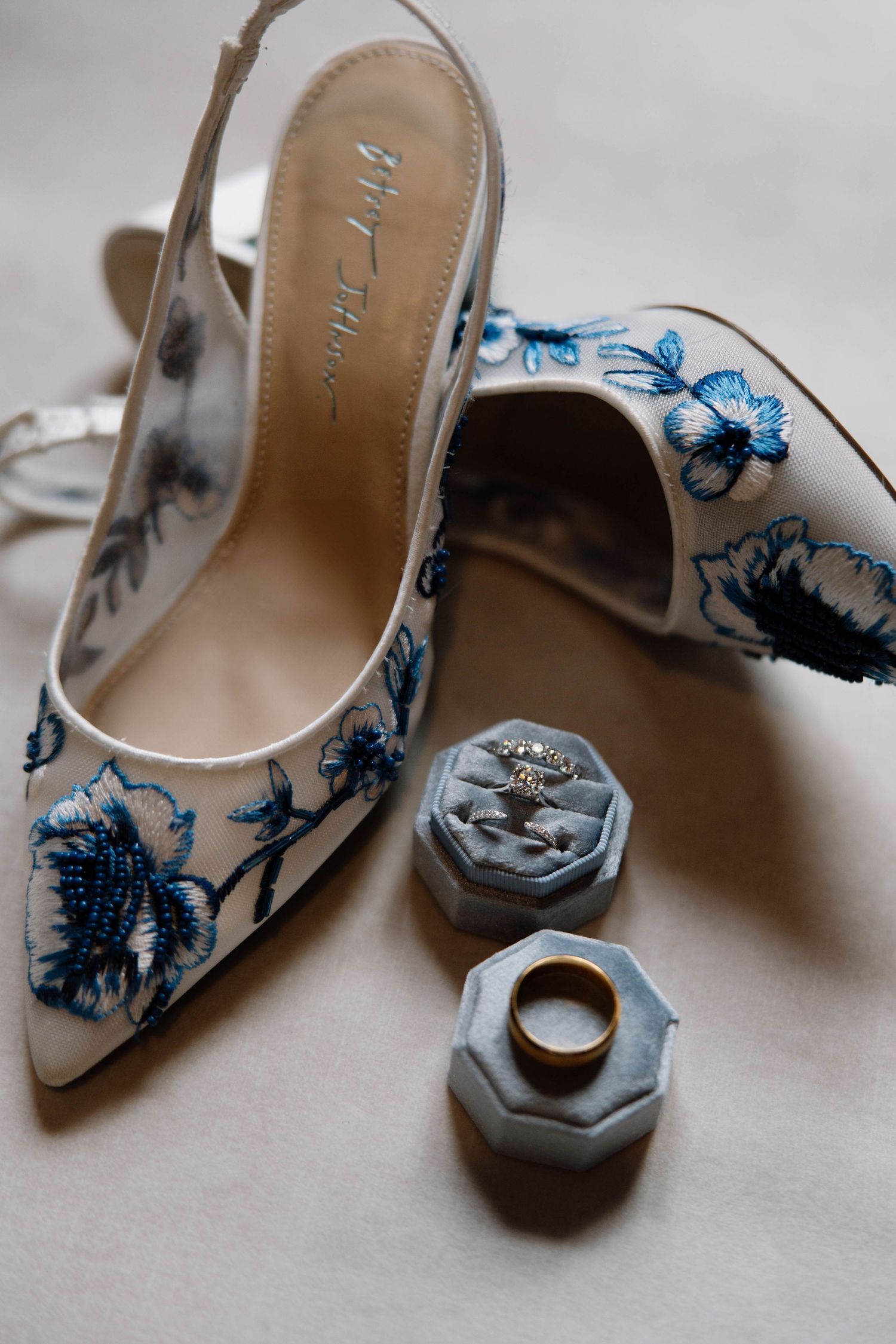 White bridal shoes with blue floral embroidery next to marble ring boxes.