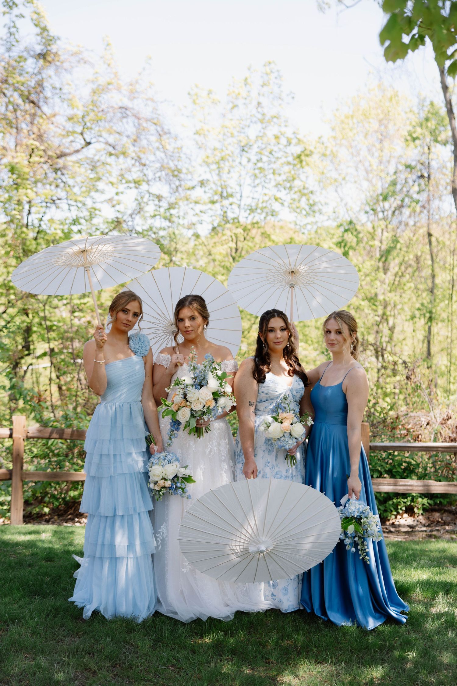 Wedding party with bridesmaids in flowing blue gowns holding decorative white parasols during outdoor photo shoot.