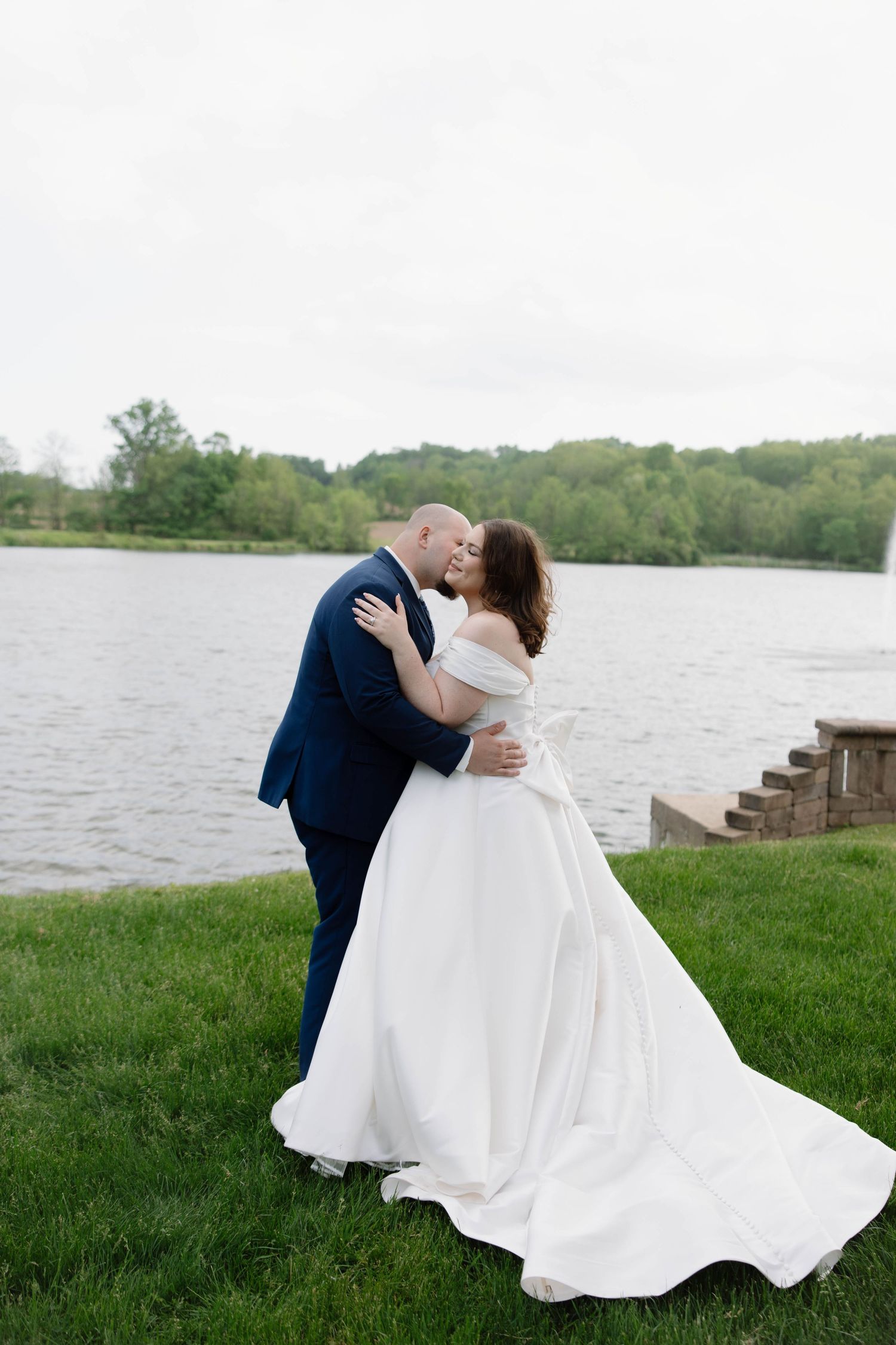 A couple in wedding attire embrace by a lake with a white gown flowing on the grass.