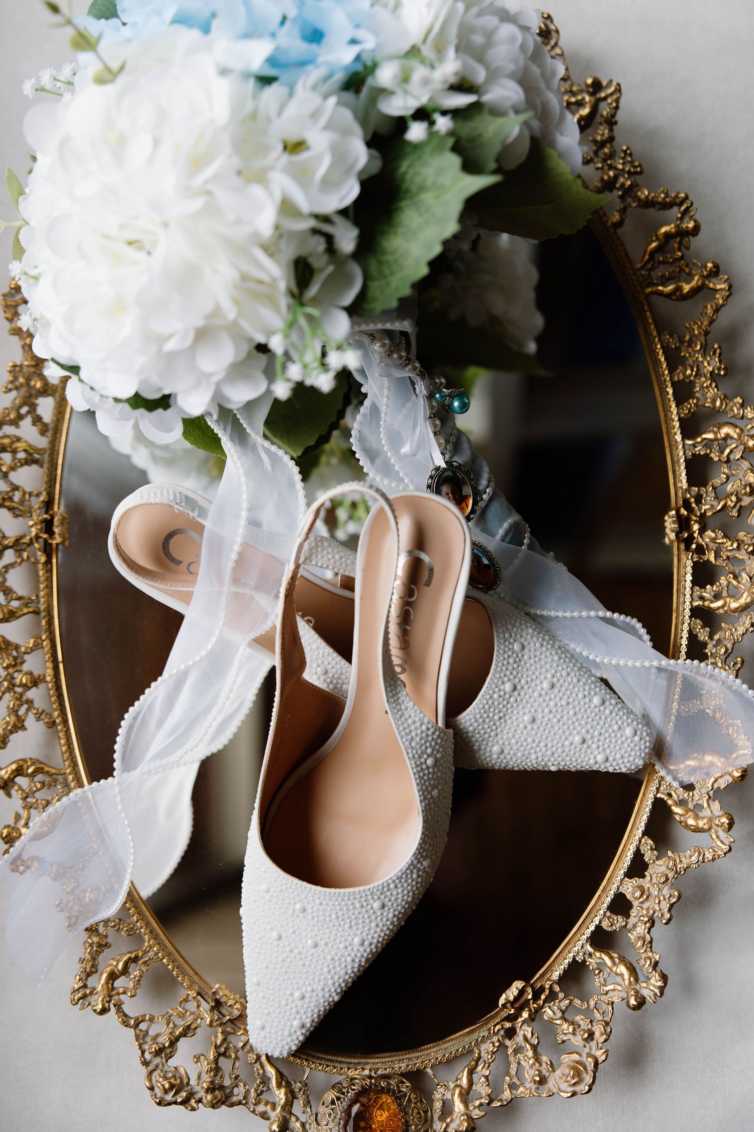 White bridal shoes and floral bouquet displayed on ornate gold mirror with intricate frame details.