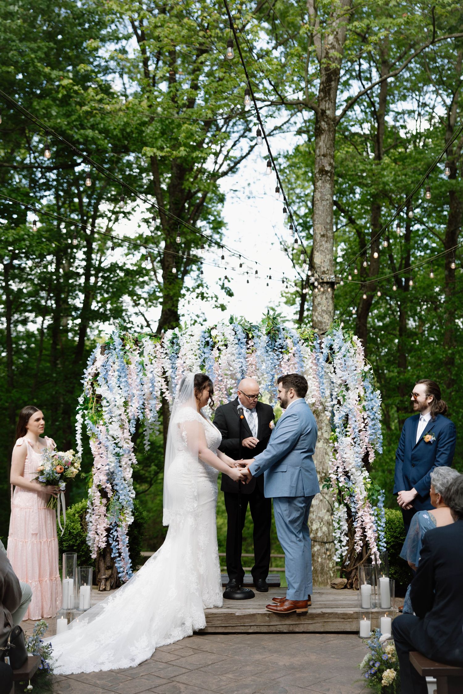 Outdoor wedding ceremony under floral arch with spring blooms and greenery in wooded setting.