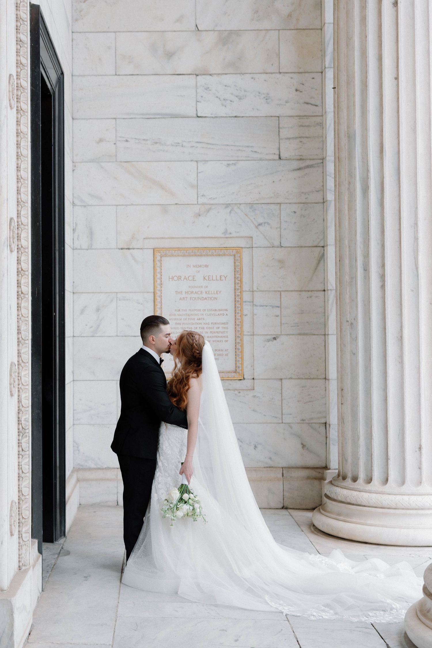 A romantic wedding couple shares intimate moments between elegant white marble columns in a classic architectural setting.