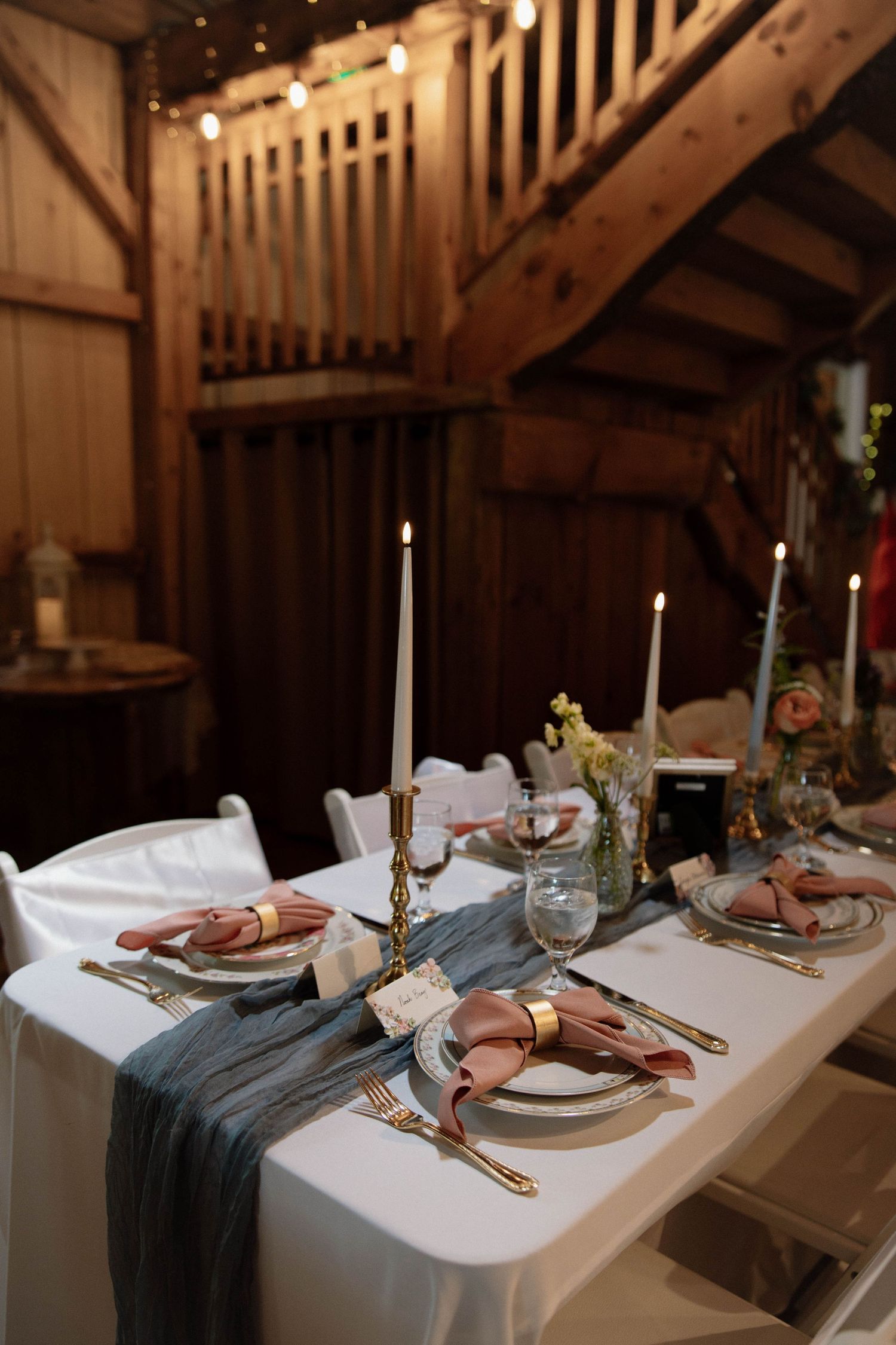 Elegant table setting at a rustic barn wedding with gold cutlery, white linens, blue runner and tall white candles.