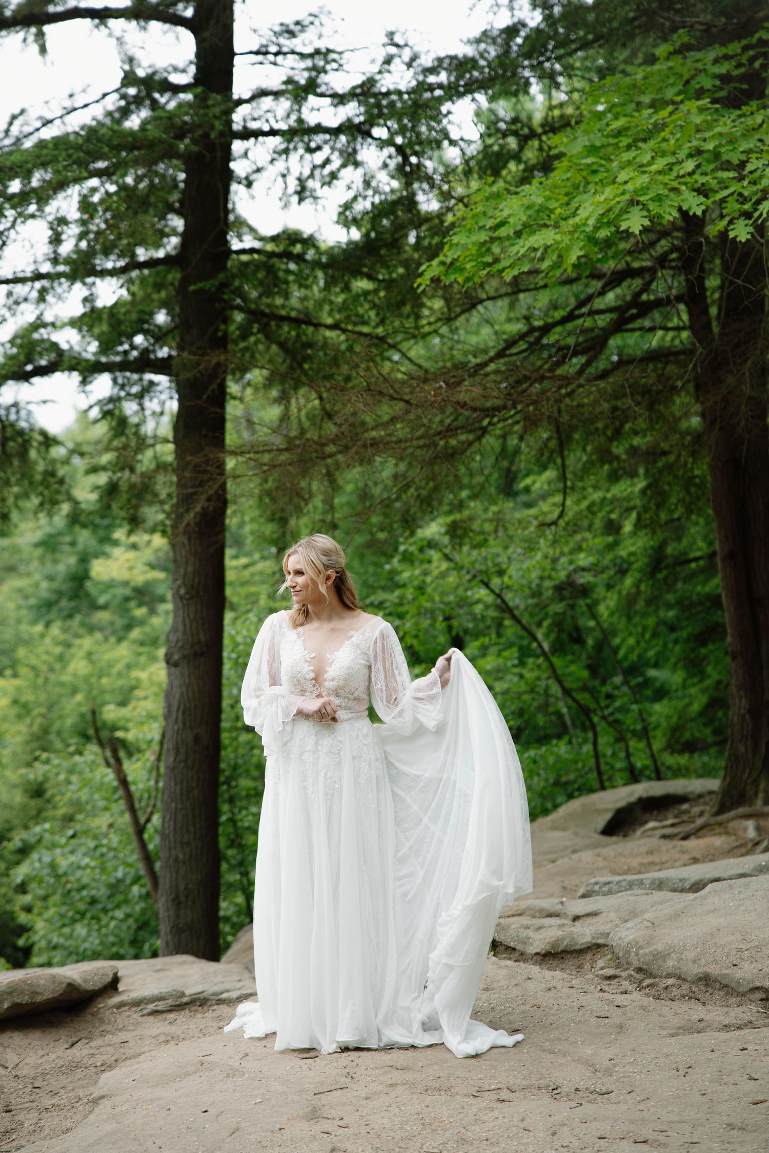 A figure in a flowing white wedding dress stands on a rocky overlook surrounded by lush evergreen forest.