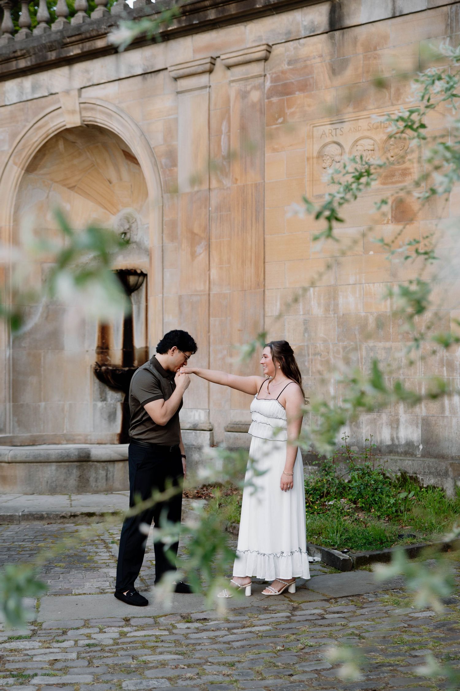 A romantic couple dancing on cobblestone pavement near a stone archway with blooming foliage in spring.