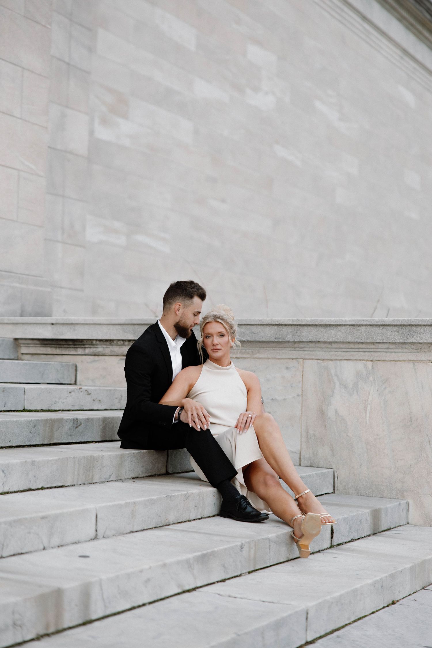 A couple in formal attire sits together on white marble steps against a light stone wall backdrop. Cleveland, OH