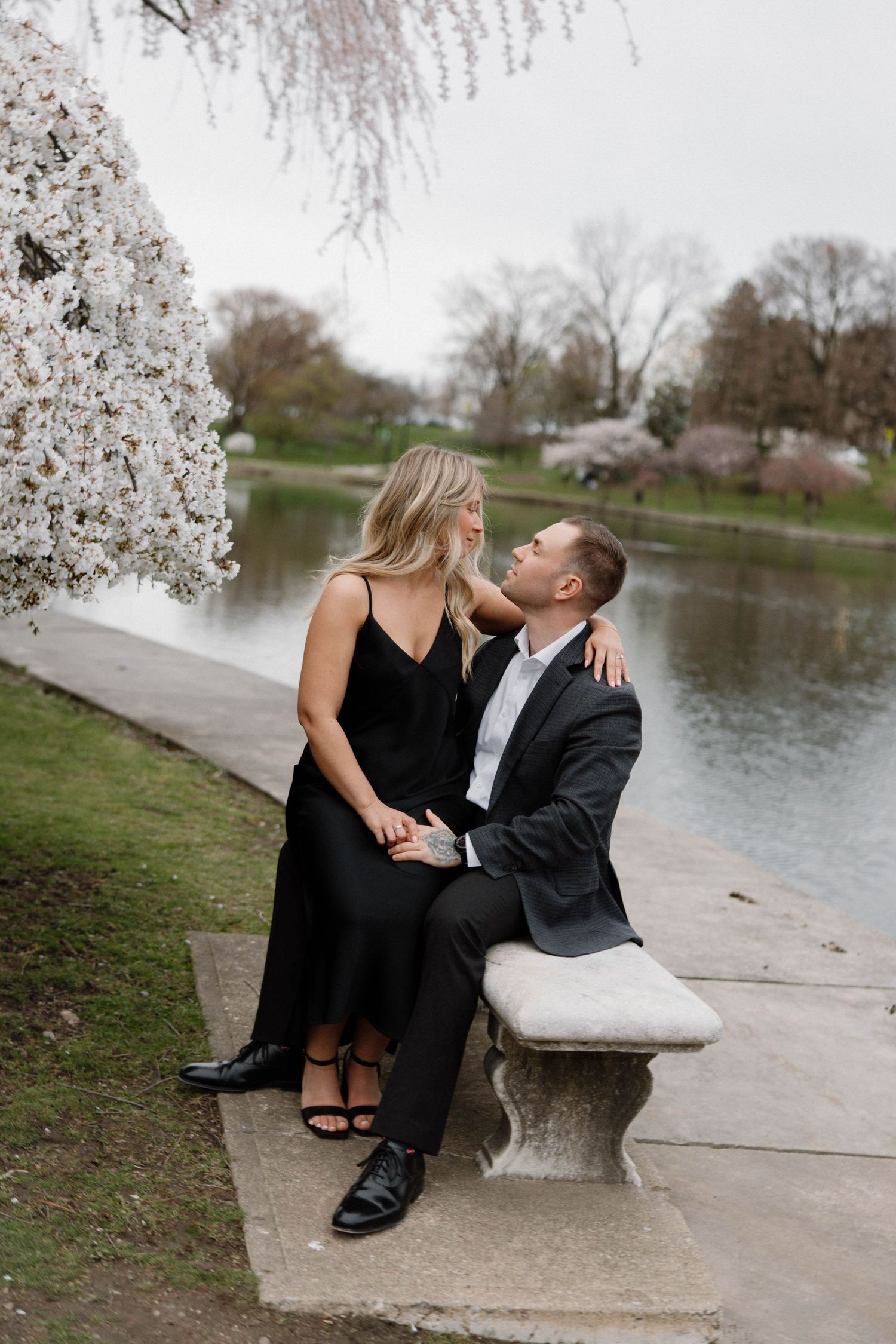 Elegant couple in formal attire sharing a romantic moment on stone bench by a lake under blooming cherry blossoms. Cleveland, OH