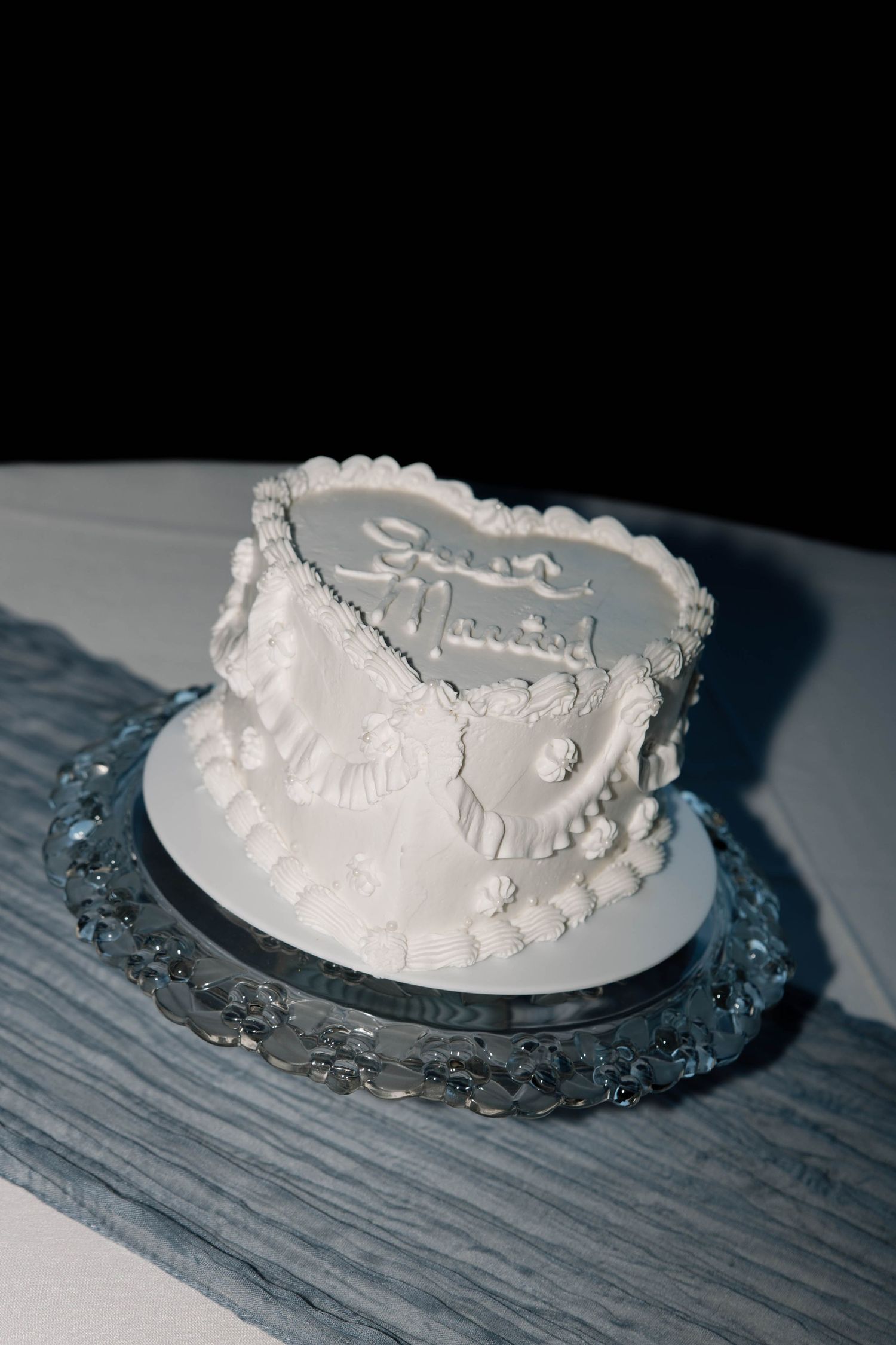 A small white wedding cake with delicate piping details sits on a glass cake stand against a dark background.