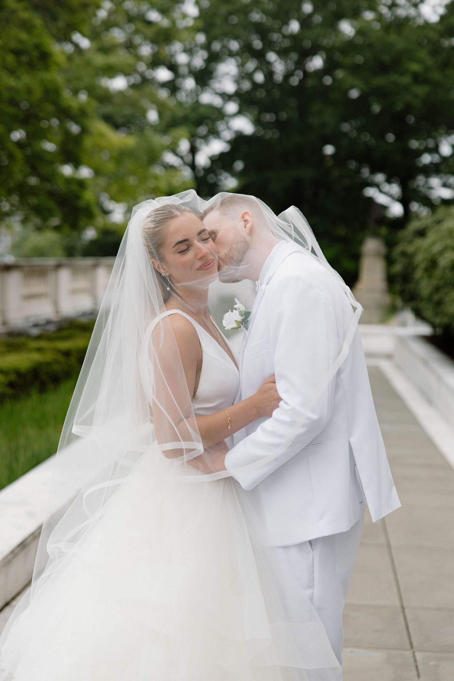 Newlyweds share intimate moment in white wedding attire with lush garden backdrop.