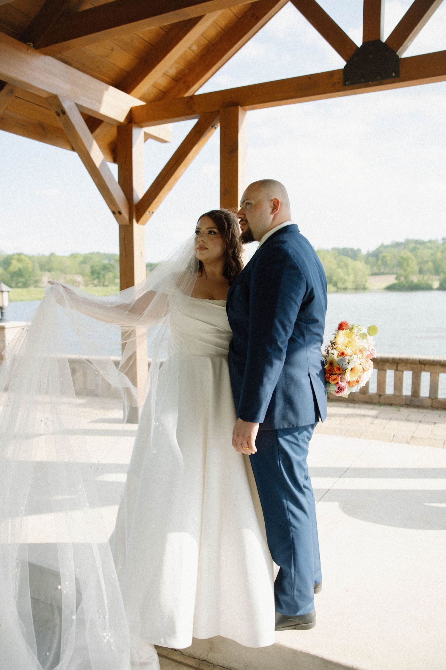 A wedding couple stands under a wooden pavilion overlooking a lake, with the bride's veil flowing in the breeze.
