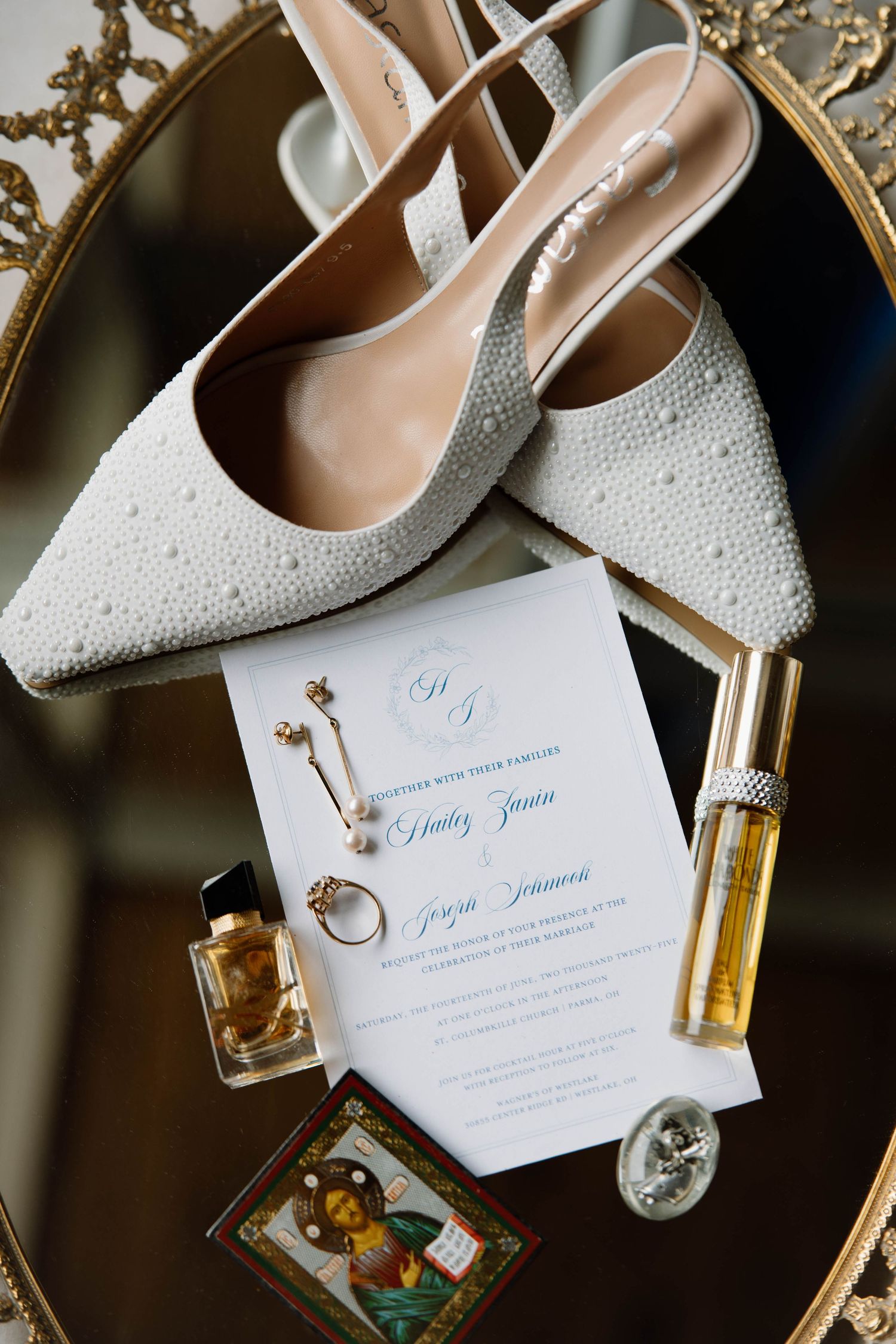 Wedding details flat lay with white beaded bridal shoes, invitation, perfume bottles and jewelry on ornate mirror.