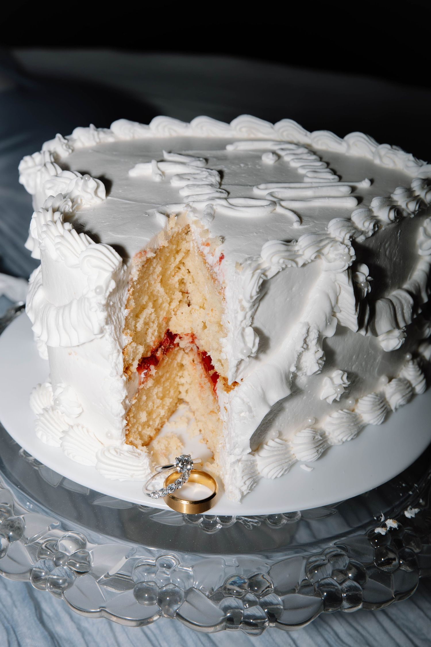 A white frosted wedding cake with a slice removed showing vanilla and raspberry layers, with a diamond ring displayed beside it.