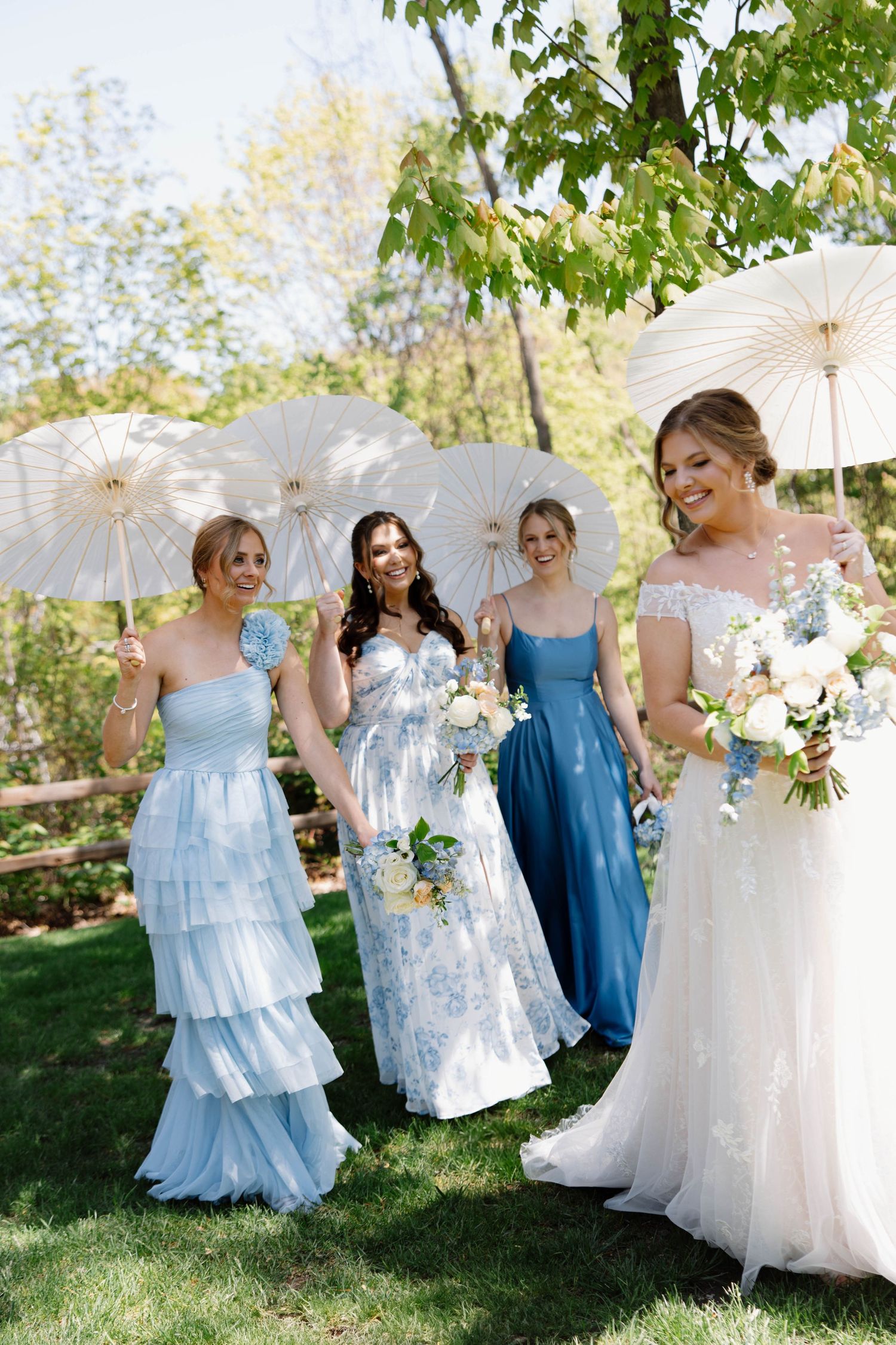 A wedding party poses outdoors with white parasols and blue dresses under trees.
