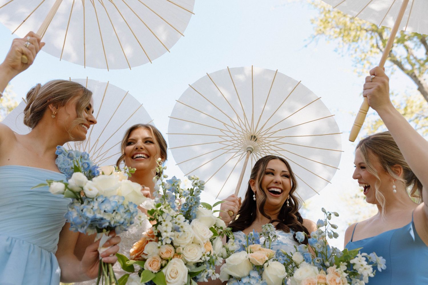A group in light blue dresses holds white parasols and floral bouquets while joyfully celebrating outdoors.
