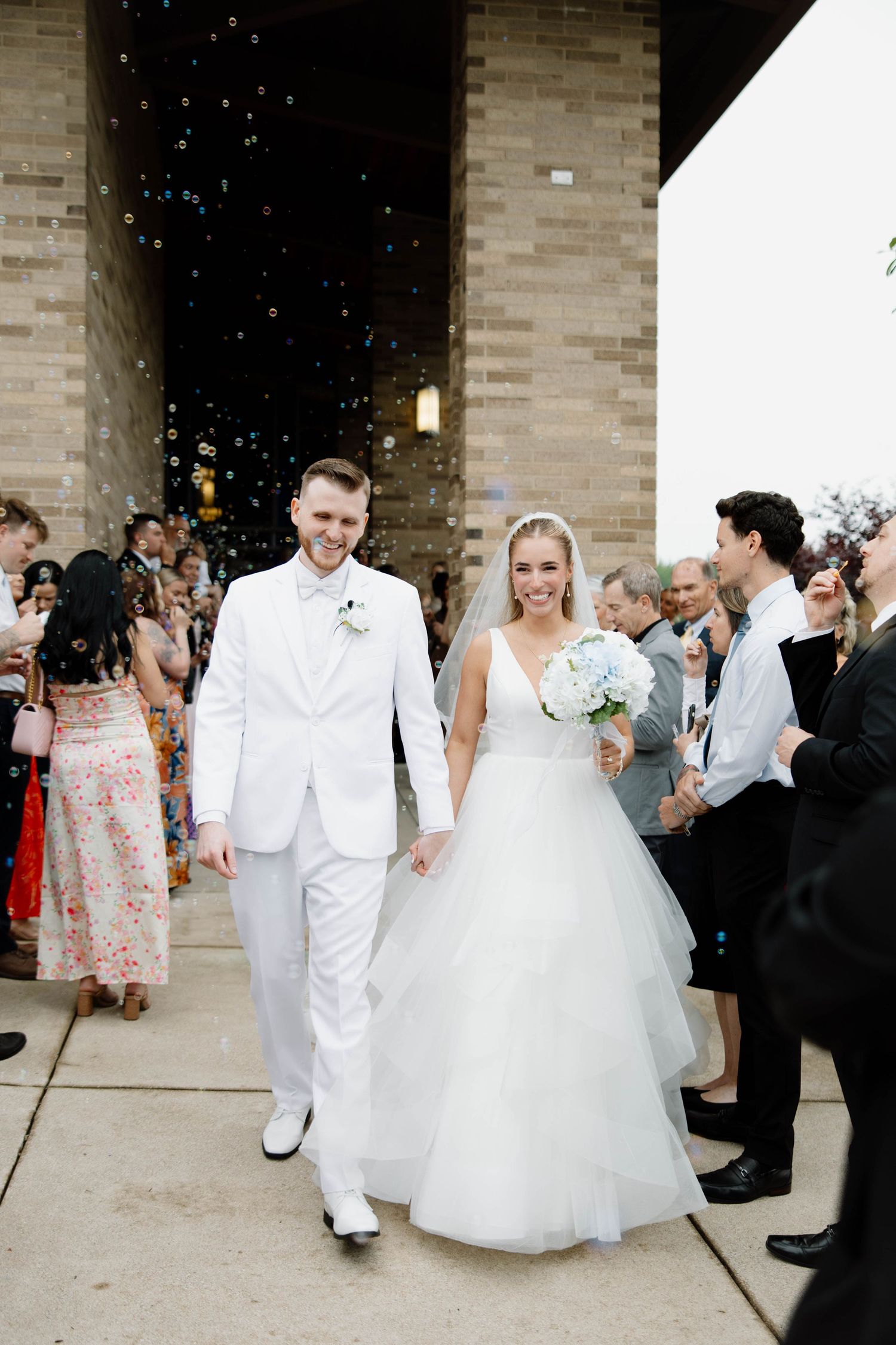 Newlywed couple exits church under shower of bubbles while wedding guests line the walkway in celebration.