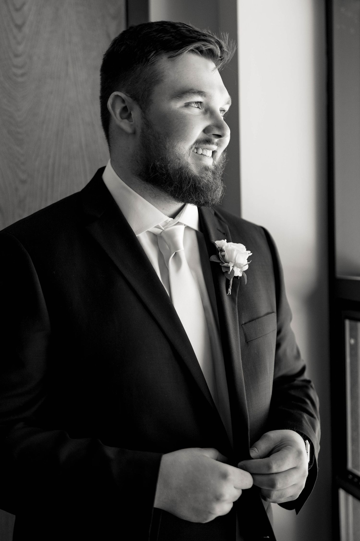 A groom in a black suit and white tie adjusts his jacket while standing near a window.