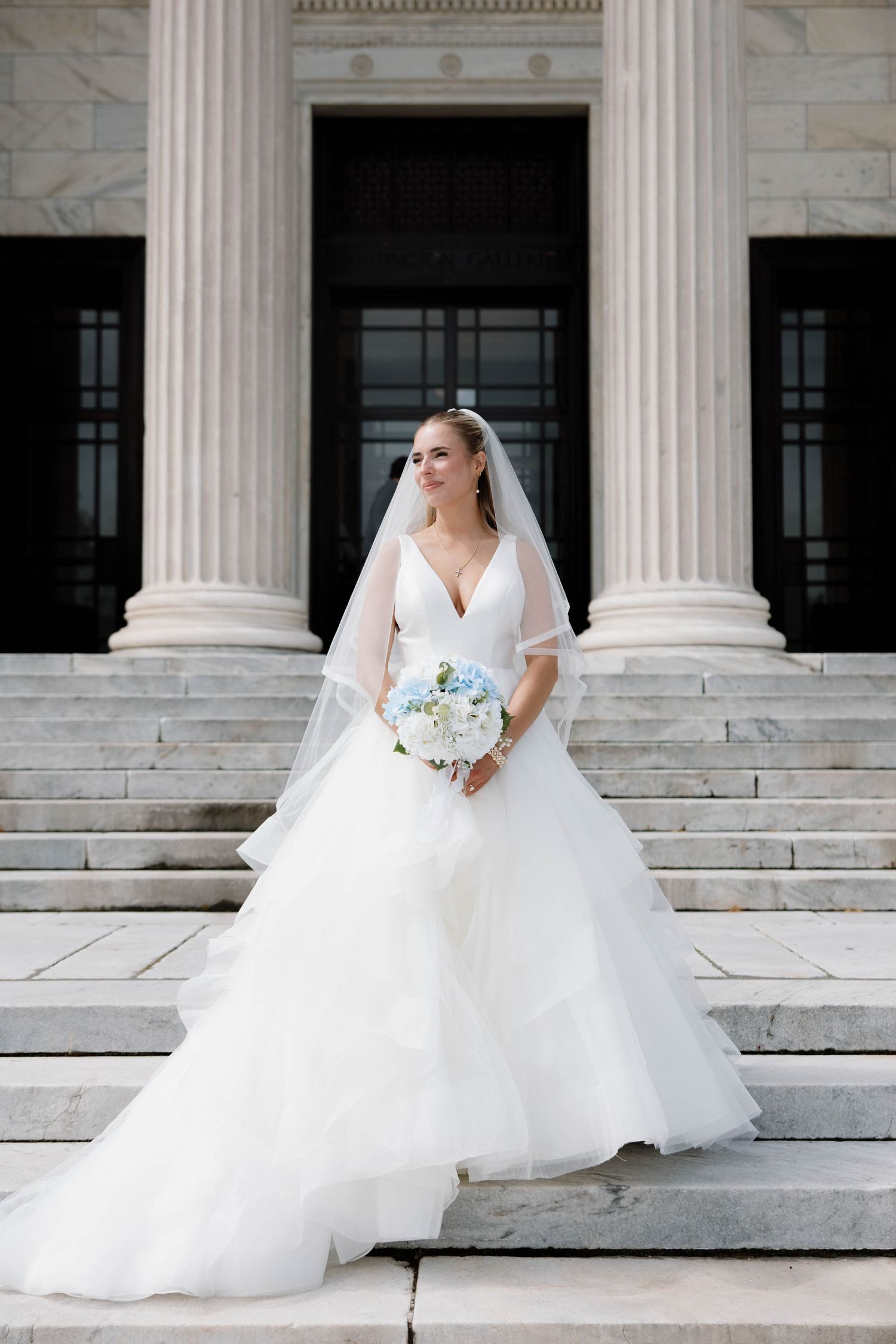 A bride in a flowing white ballgown poses elegantly on marble steps with classical columns.