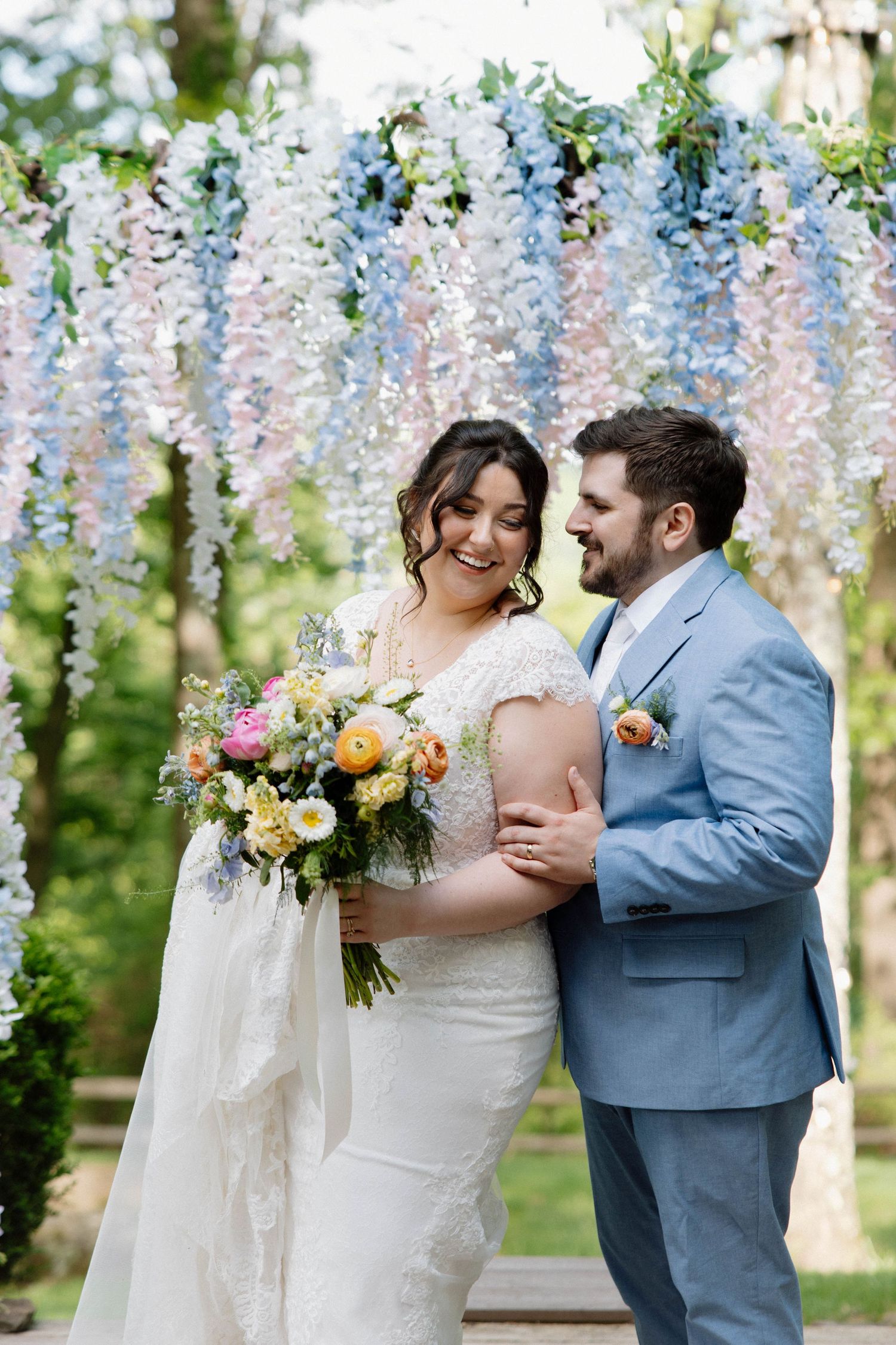 A romantic wedding couple stands under a canopy of hanging blue and white wisteria flowers in an outdoor garden setting.