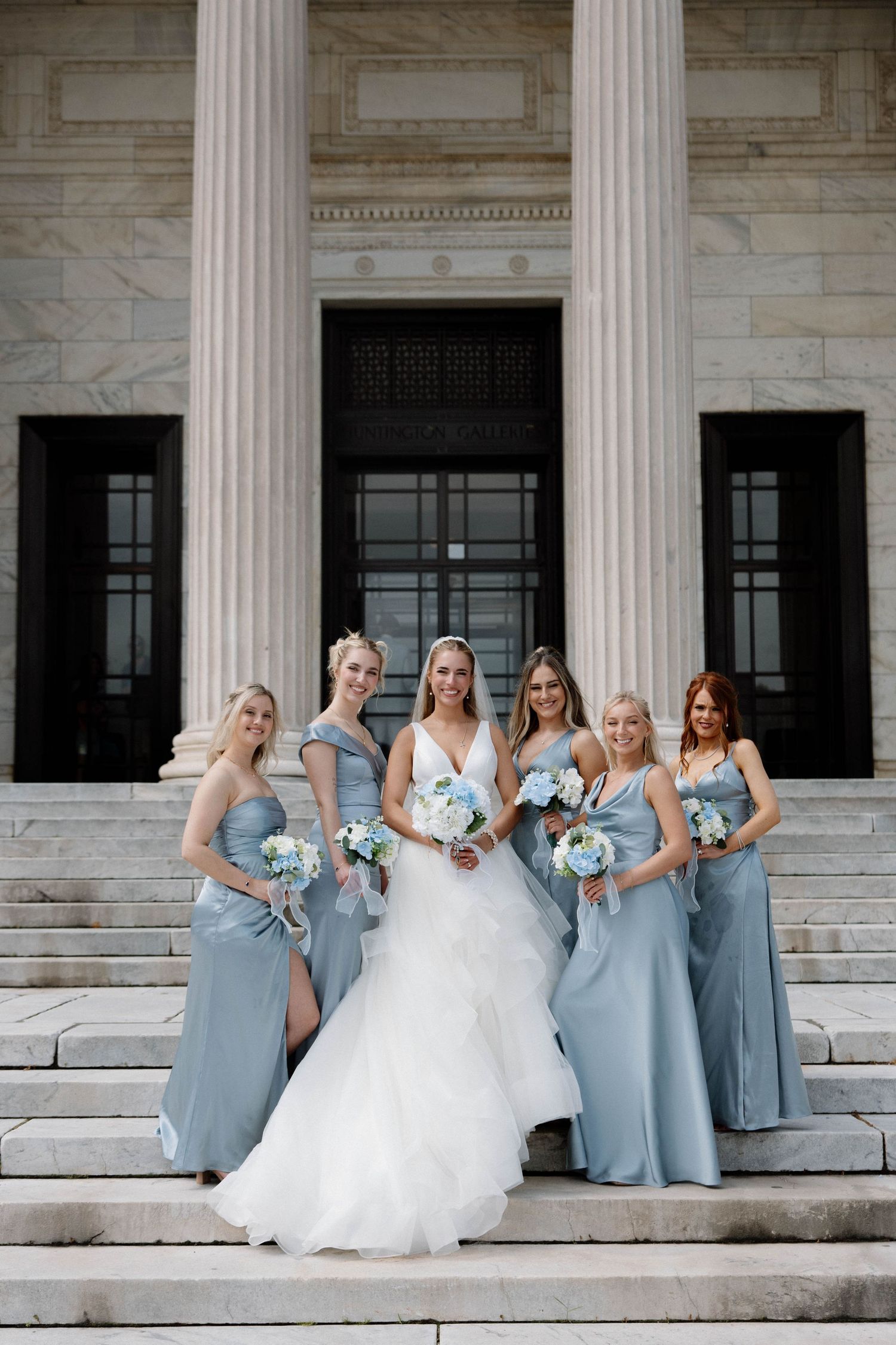 Wedding party poses on marble steps with bride in white dress and bridesmaids in light blue gowns holding bouquets.