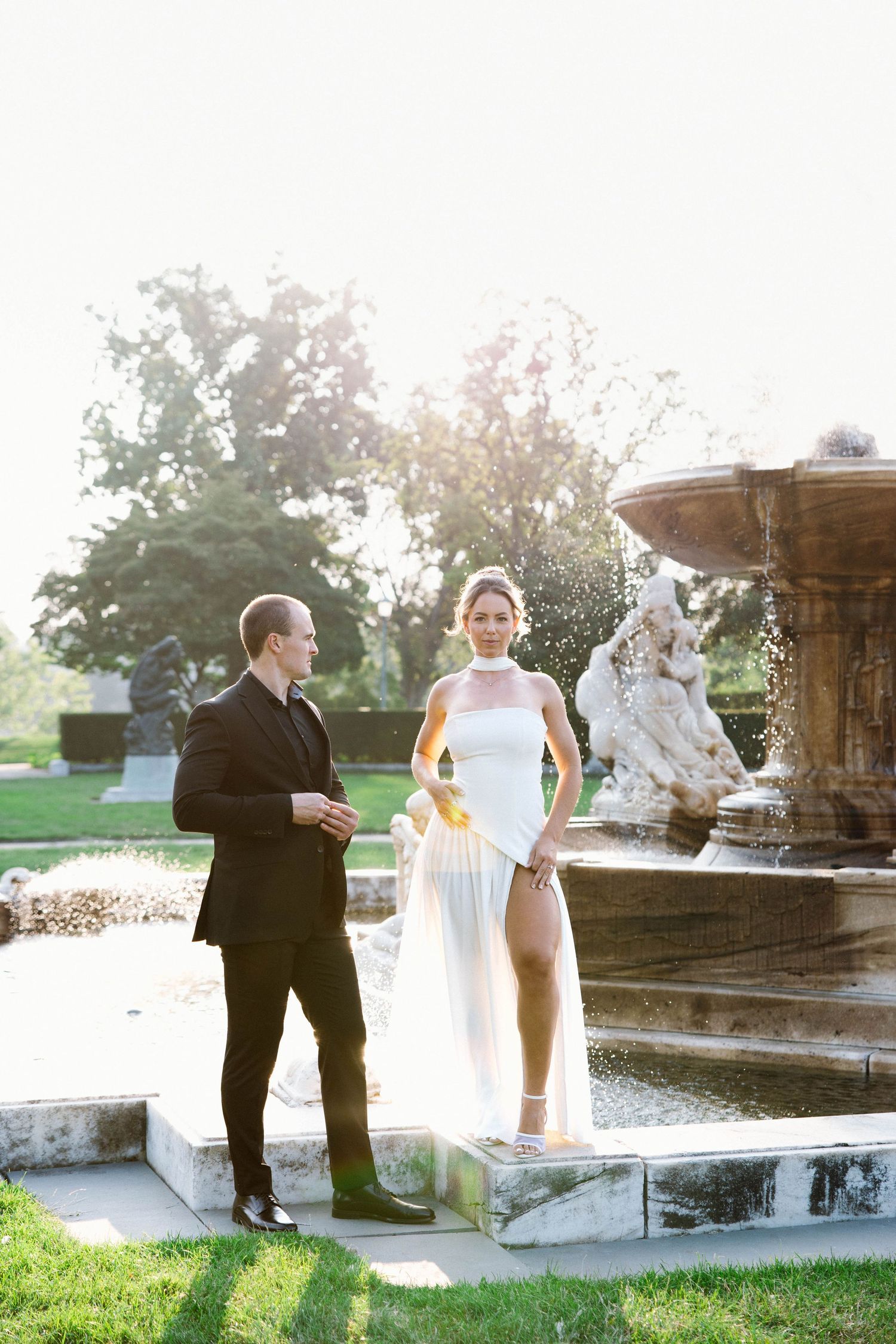 Wedding couple stands near ornate fountain in garden during romantic outdoor photoshoot at sunset.