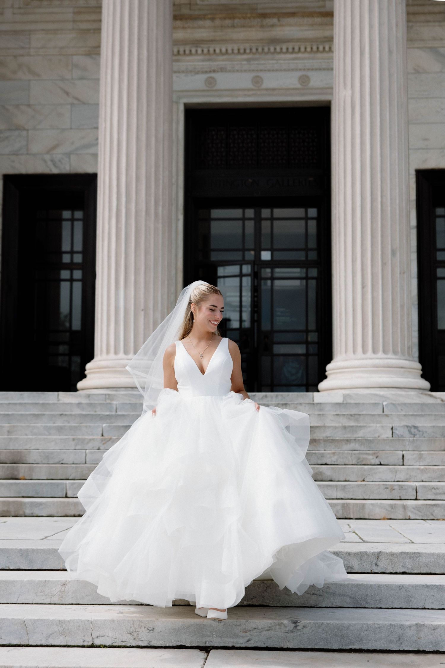 A bride in a flowing white ballgown twirls on marble steps between classical stone columns.