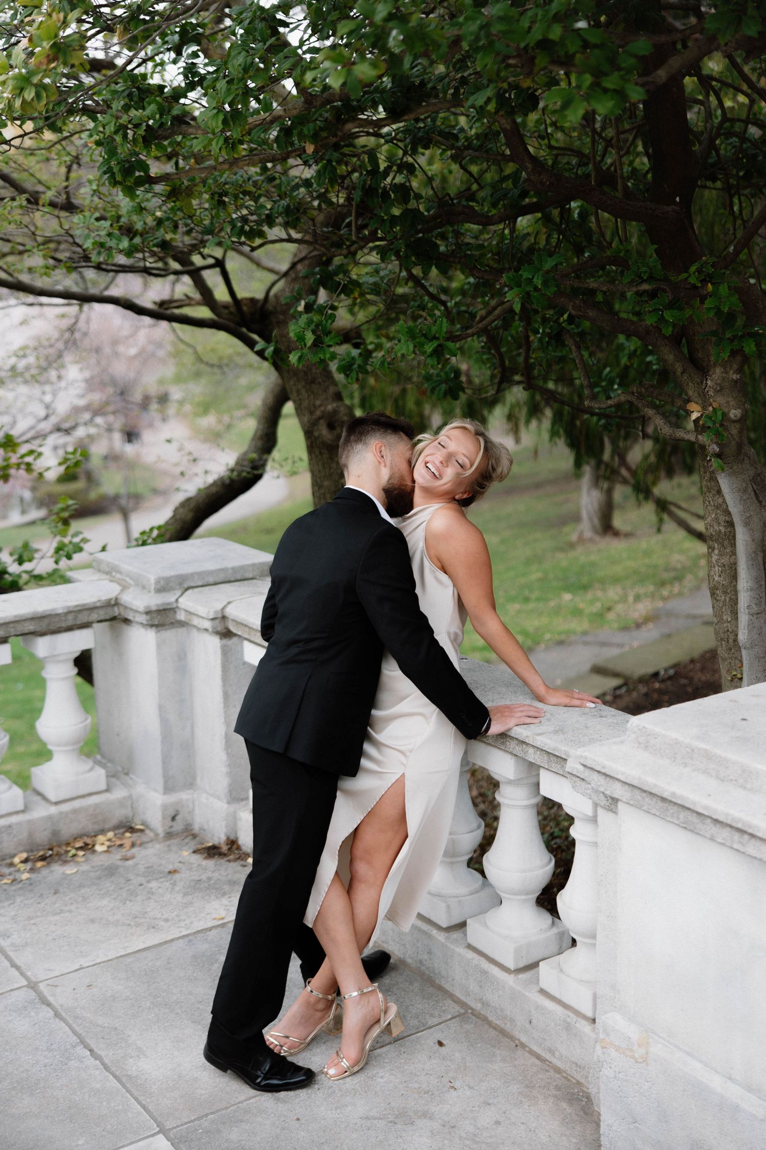 Couple in formal attire embracing on white stone balcony with trees in background. Cleveland, OH