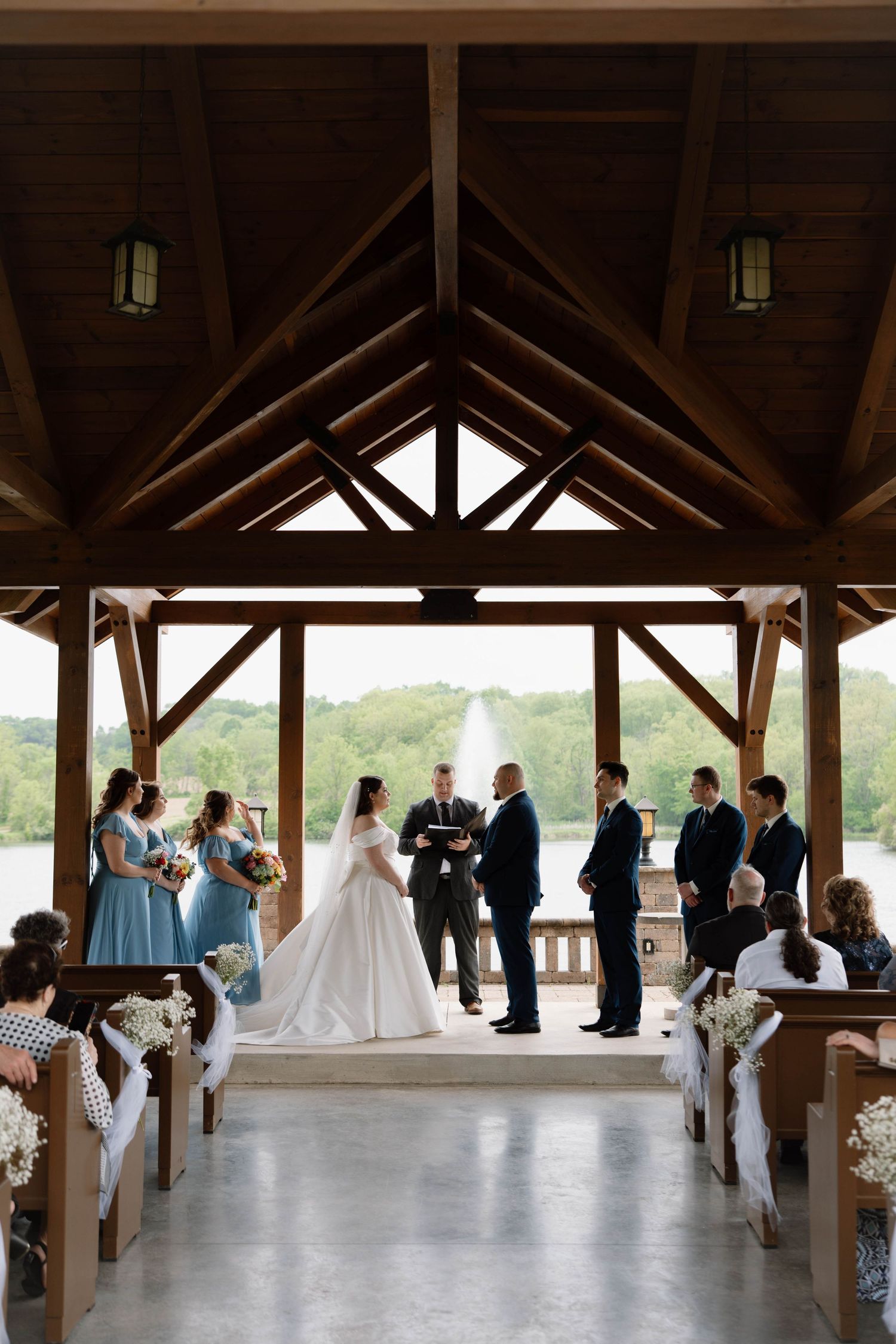 Wedding ceremony taking place in a rustic wooden pavilion with exposed beams and lanterns overlooking scenic landscape.