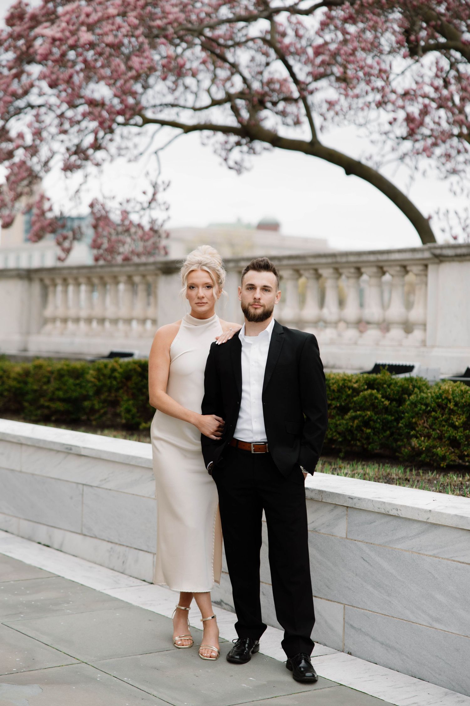 Couple in elegant attire stands beneath blooming cherry blossoms by stone balustrade at formal venue. Cleveland, OH