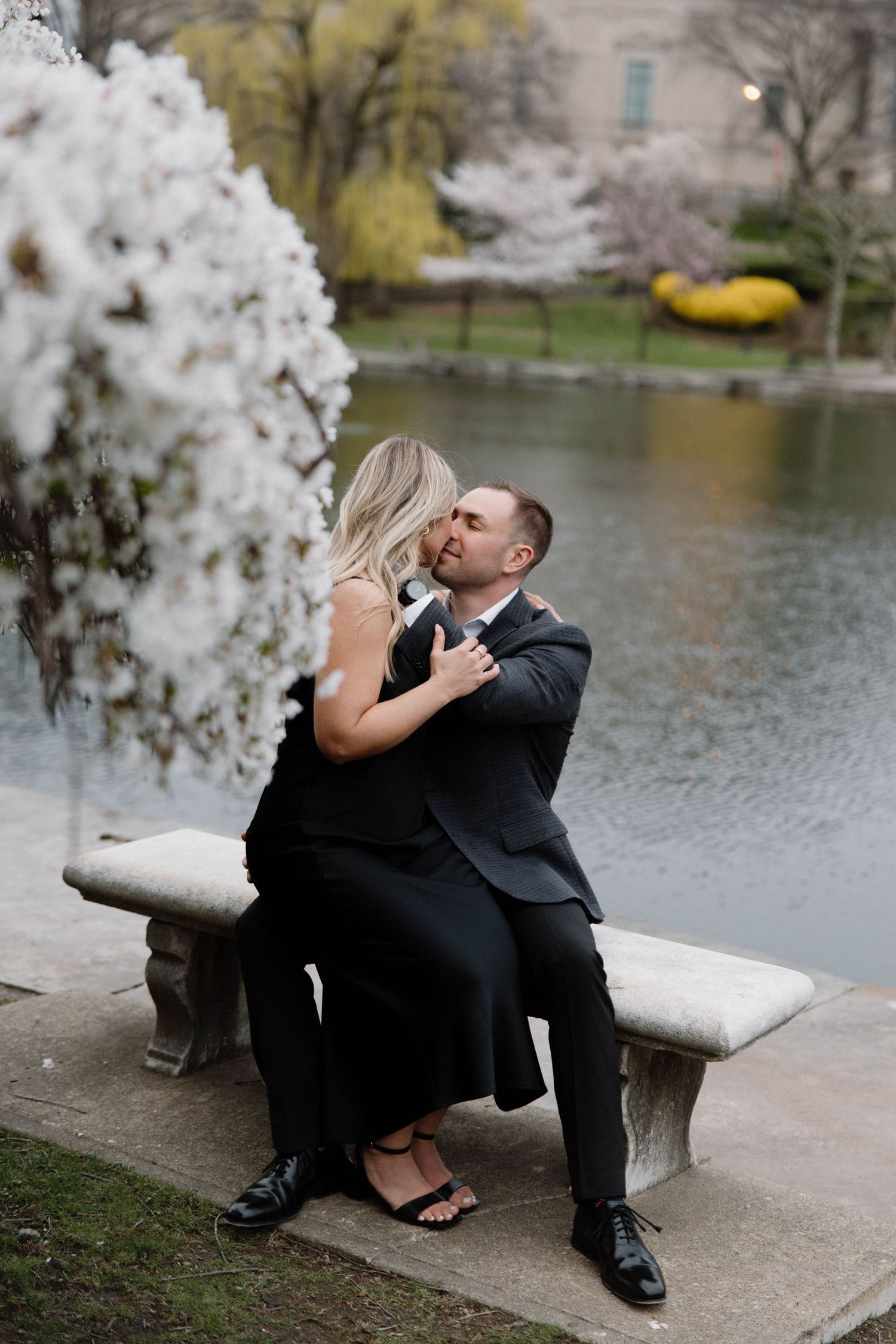 A romantic couple shares a tender moment on a stone bench by a lake with blooming cherry blossoms in spring. Cleveland, OH
