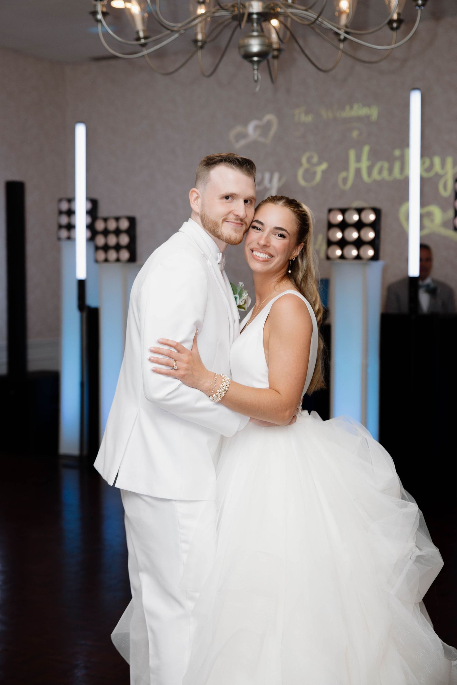 Newlyweds share romantic embrace on dance floor with chandelier and mood lighting.