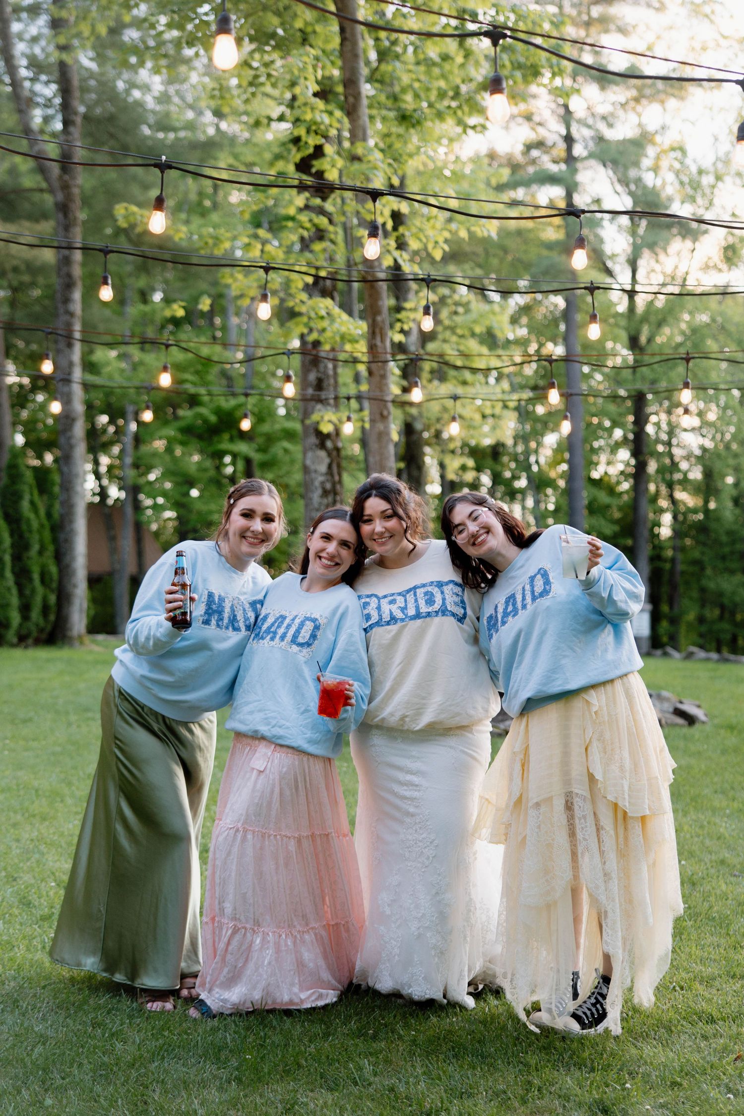 Bridal party wearing matching blue sweaters and flowing skirts pose outdoors under string lights at dusk.