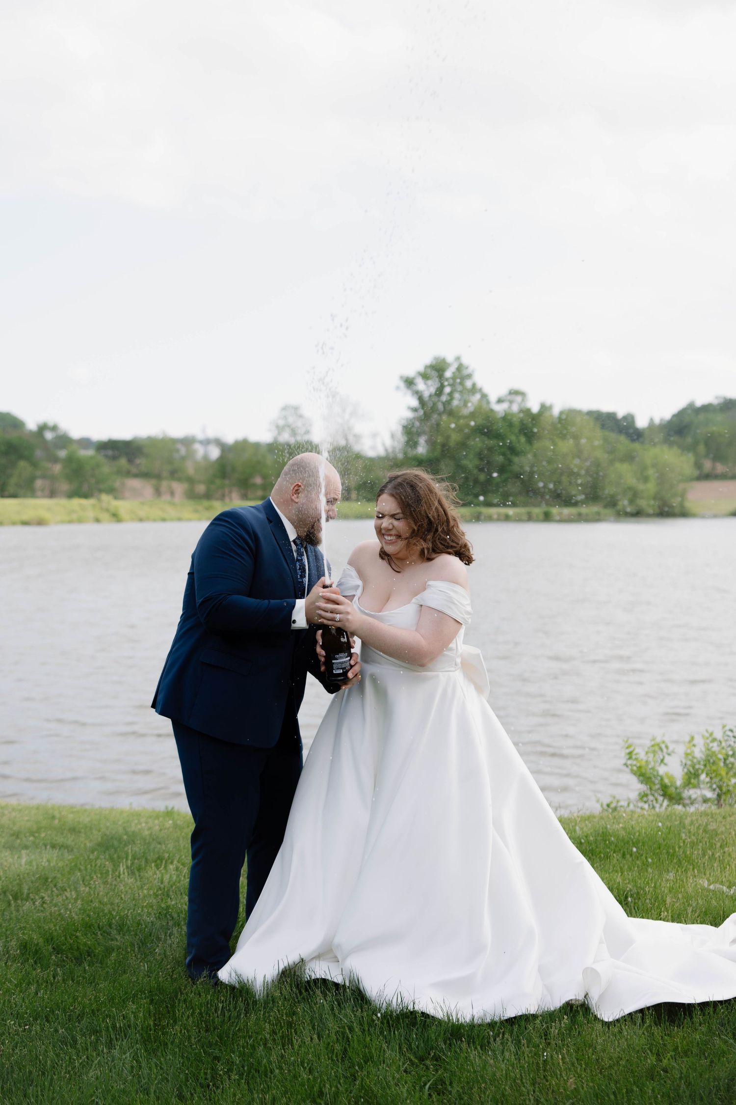 Newlyweds share an intimate moment while holding champagne glasses by a scenic lake on their wedding day.