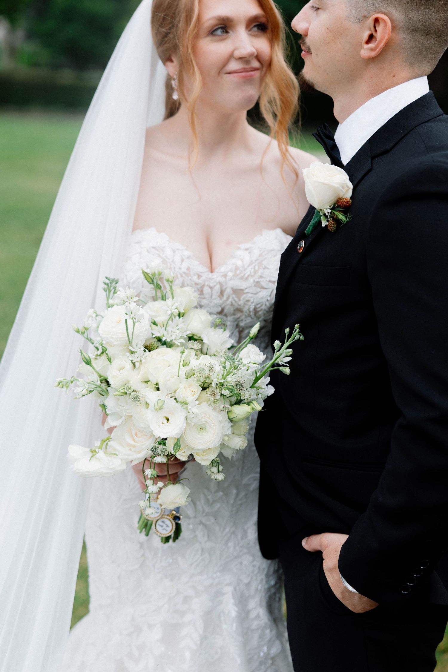 A bride in a white lace dress holding a delicate white floral bouquet poses with her groom outdoors.