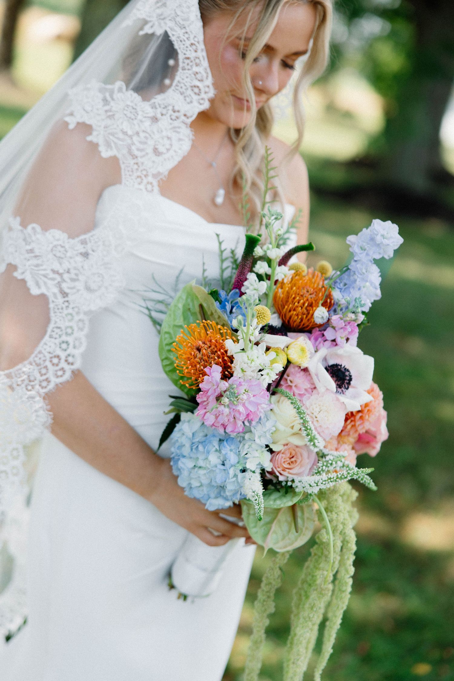 Colorful wildflower bridal bouquet featuring blue hydrangeas, orange proteas, and pink blooms with delicate lace details.