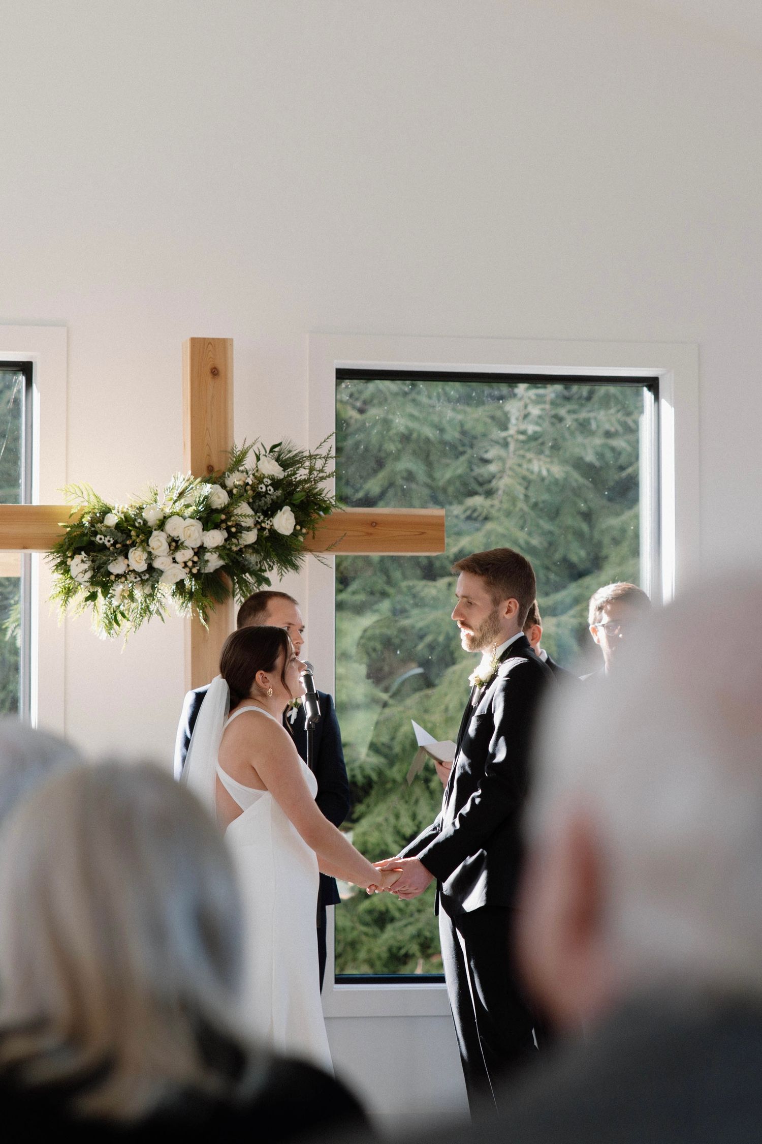 Wedding ceremony in a modern chapel with white floral arrangements and large windows.