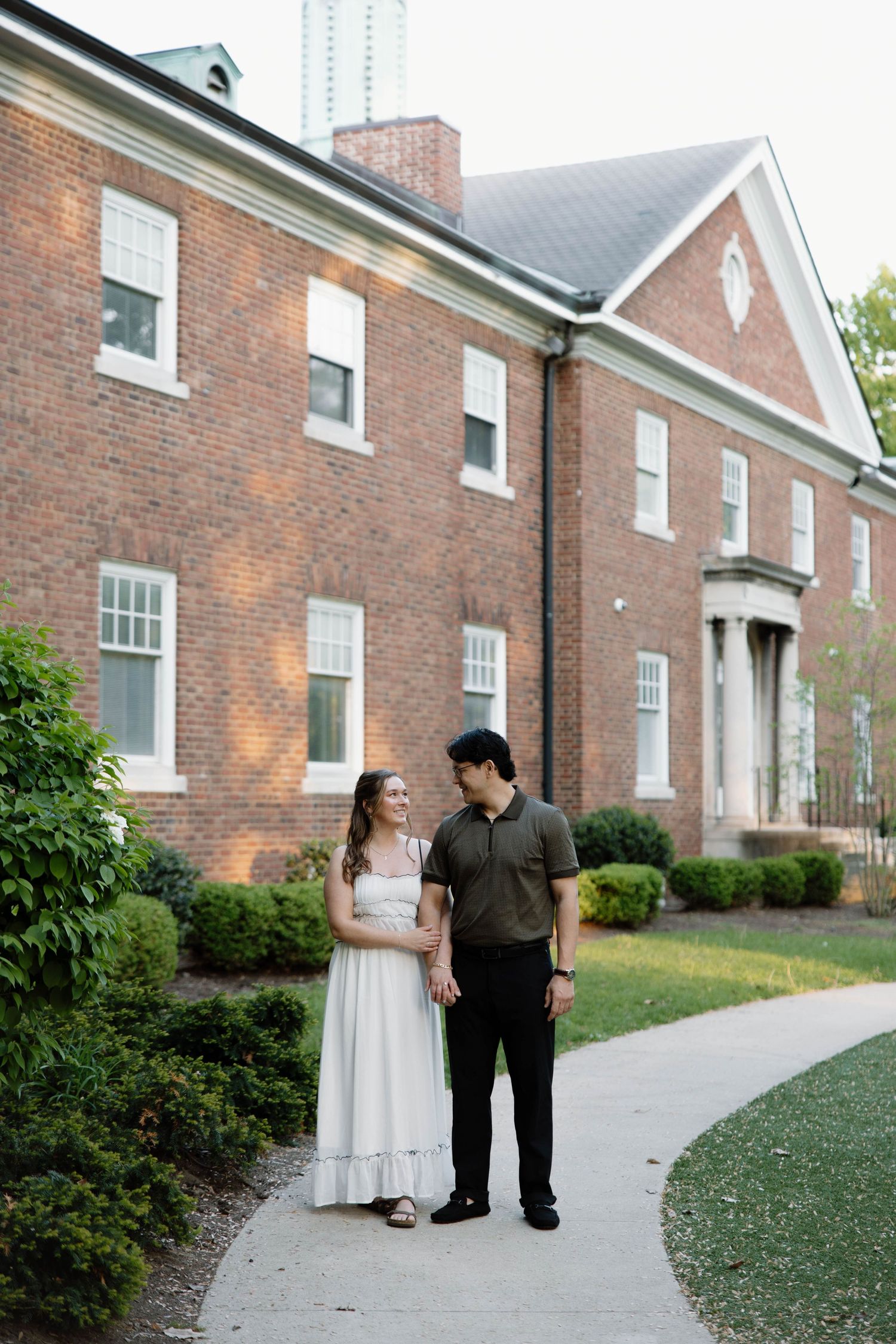 A couple stands together along a sidewalk outside a grand red brick colonial style building on a summer day.