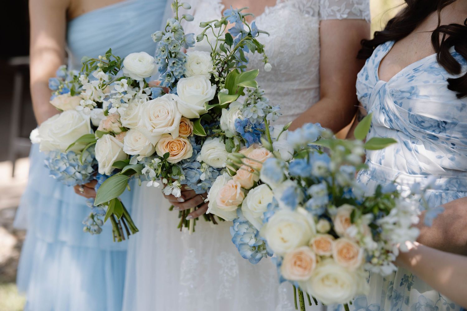 Wedding party holds beautiful pastel bouquets with white roses and light blue delphinium flowers against blue dresses.