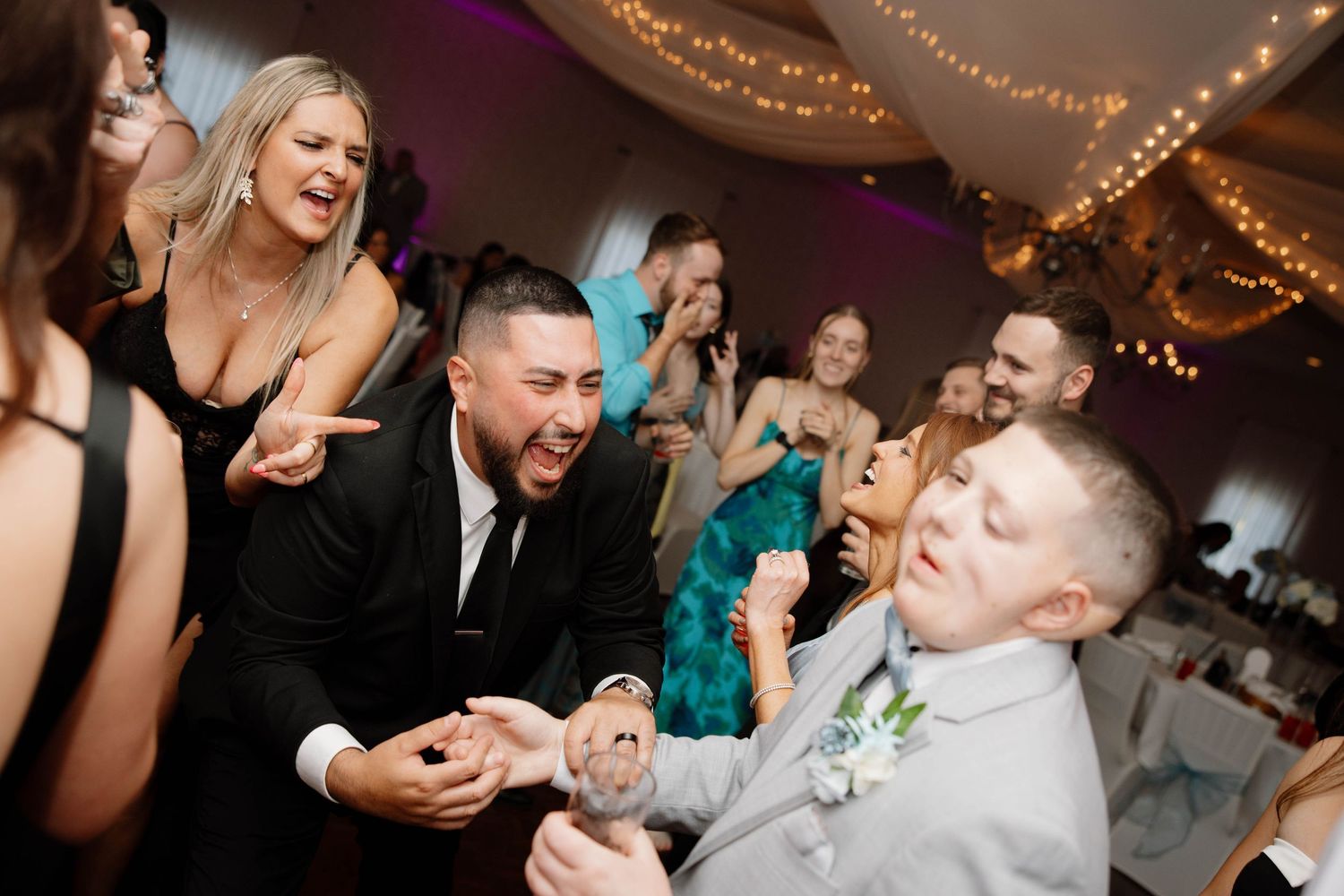 A festive wedding venue features string lights overhead while guests celebrate and dance on the dance floor during a reception.