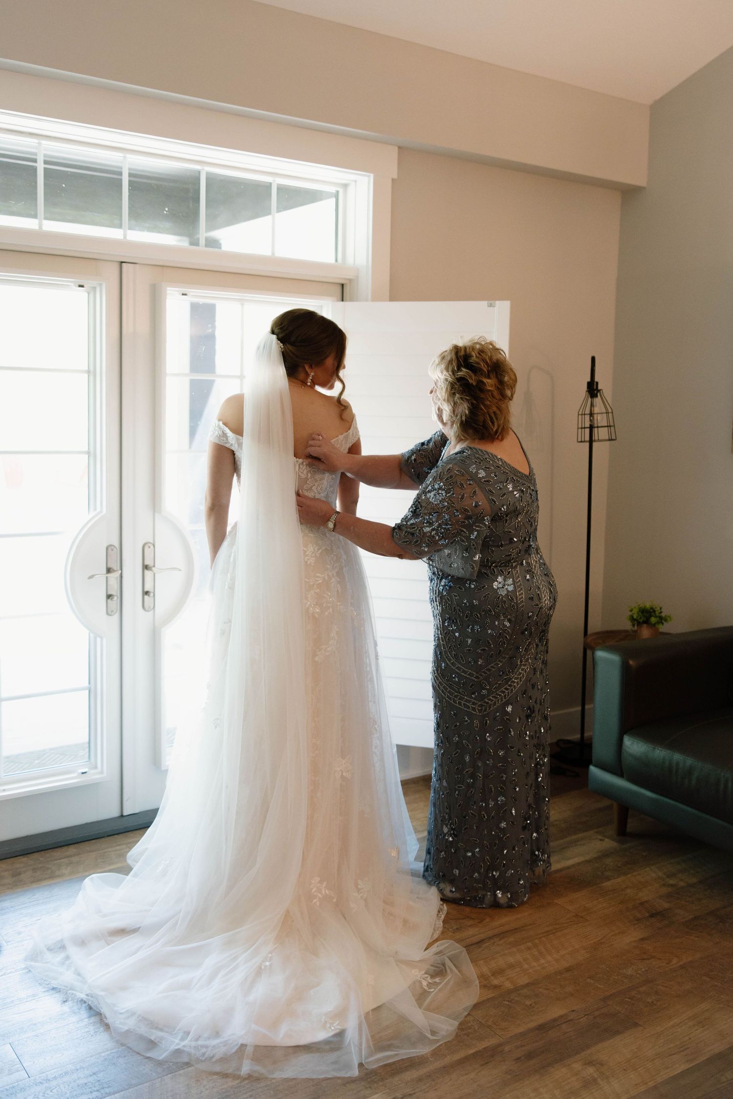A flowing white wedding dress being adjusted in a sunlit room with hardwood floors.