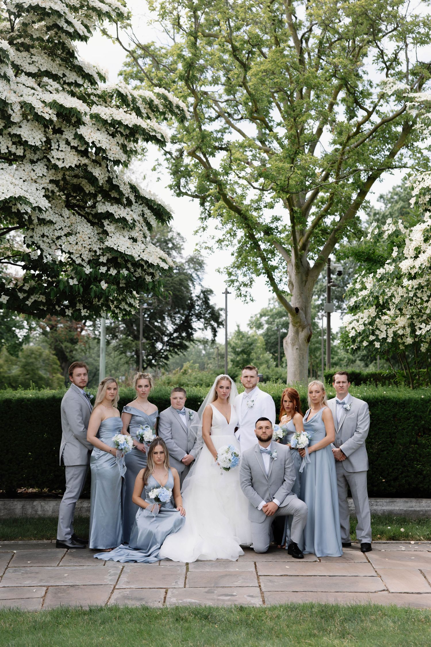 Wedding party in gray and white attire poses together on stone steps with lush greenery backdrop.